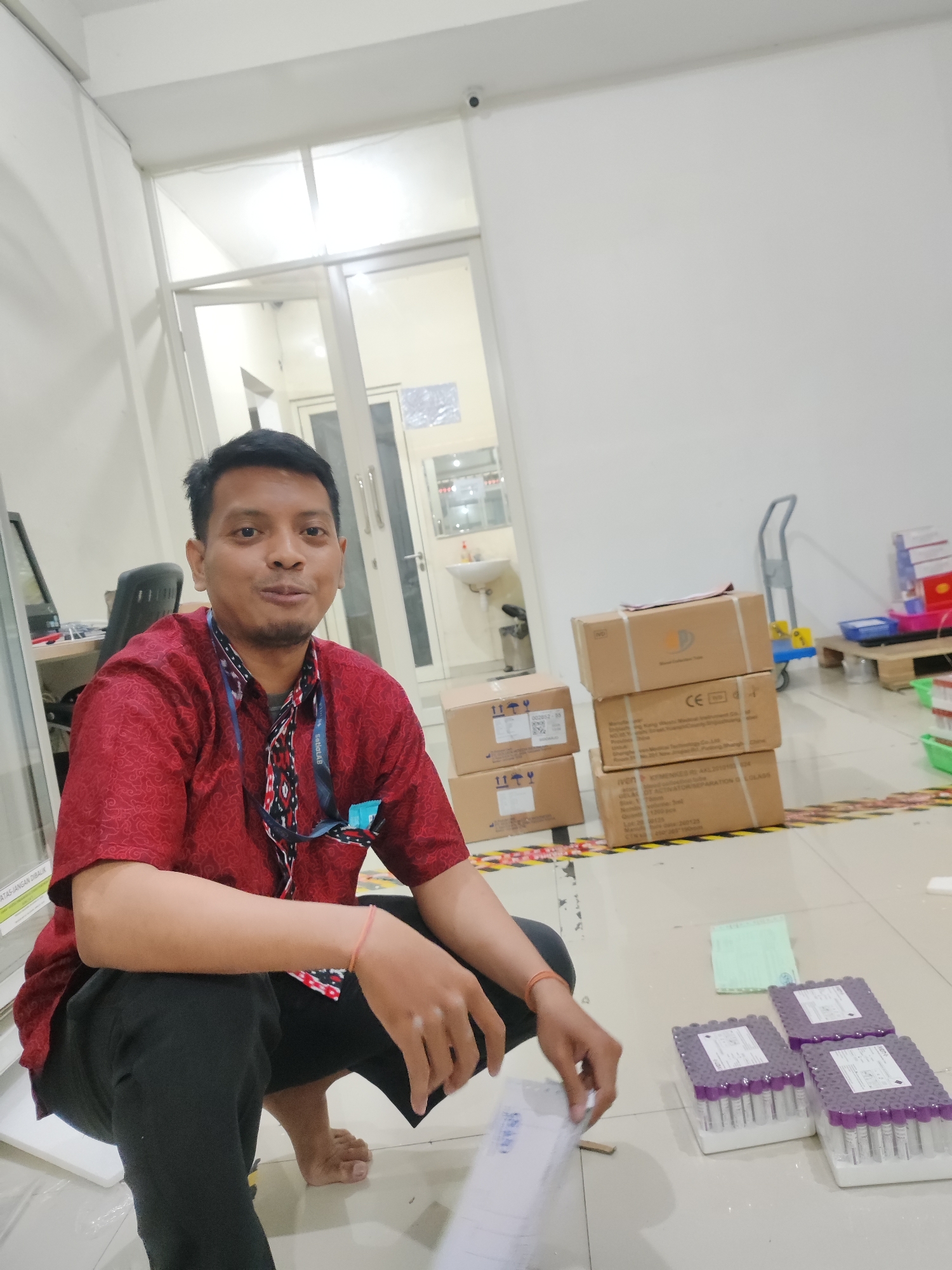 A man wearing a red shirt and black pants kneels on the floor in what appears to be a medical facility. He is holding a white package and looking towards the camera. Behind him, there are several cardboard boxes stacked on top of each other. To his right, there are racks of purple-capped vials, possibly medical samples. In the background, there is a door with glass panels, a sink, and a rolling cart. The floor is tiled and there is caution tape visible. The overall impression is a work environment, possibly a laboratory or storage area, in Pepelegi, Indonesia.