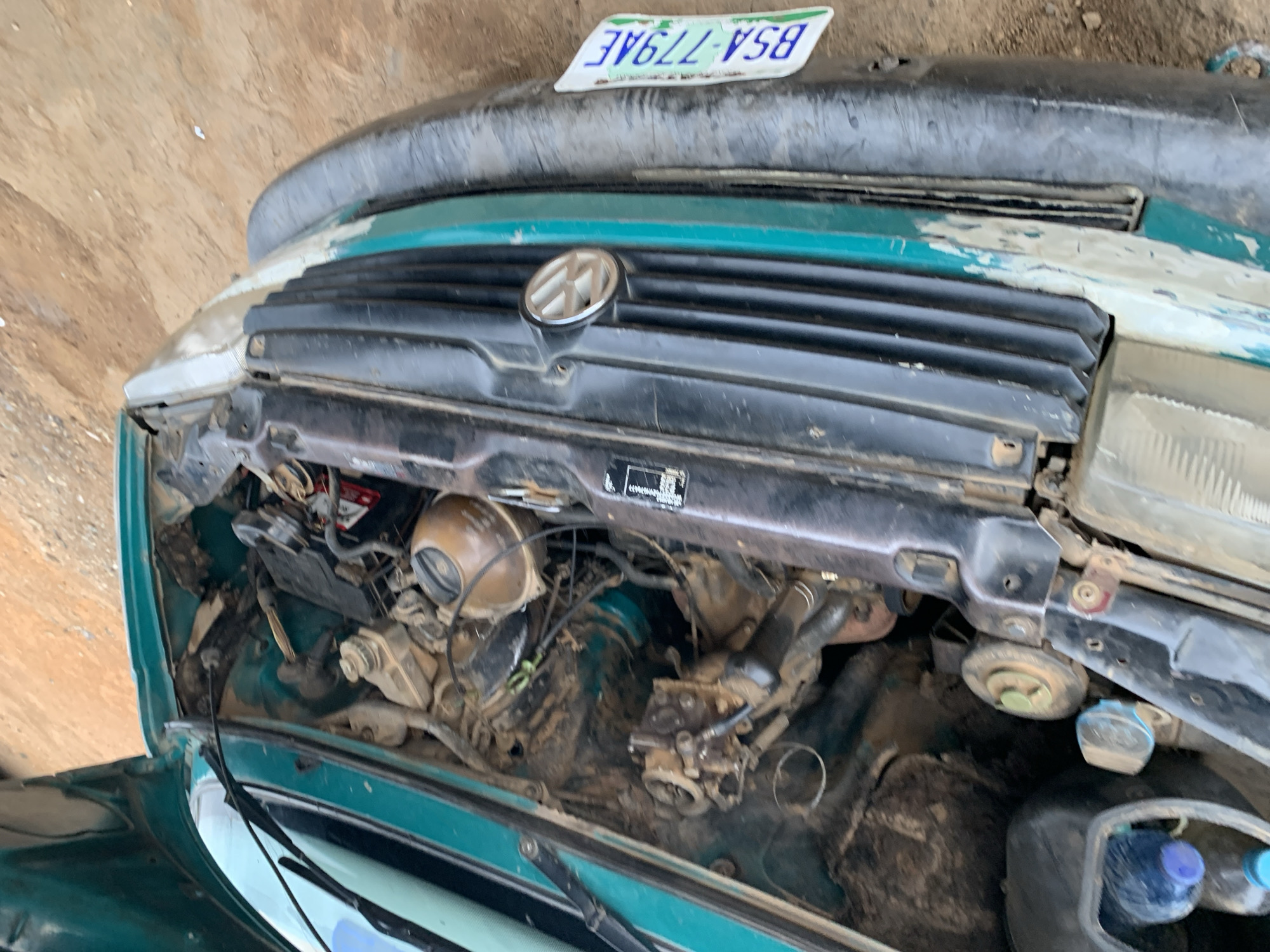 The image depicts the open hood of a Volkswagen car in what appears to be an outdoor location. The car is green with a black grill featuring the Volkswagen logo. The engine compartment is visible, filled with various engine components, wires, and fluids, and covered with dirt. The license plate reads "BSA-779AE". Based on the provided location, this scene is within Rafin Par, Nigeria.