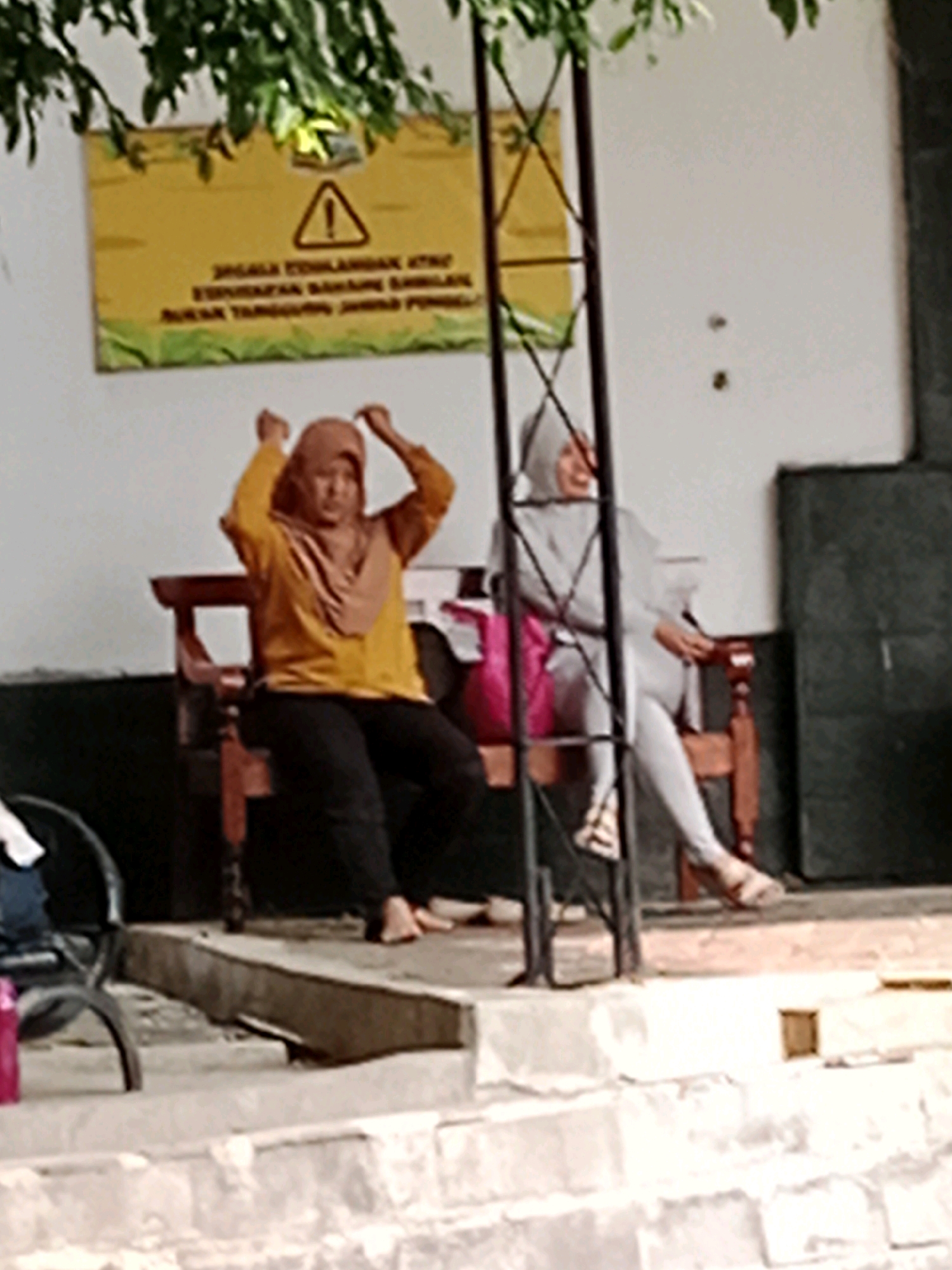 The image depicts an outdoor scene with two individuals seated on a wooden bench. The individual on the left is wearing a yellow shirt and a light brown hijab, with black pants. They are in a seated position with their arms raised and hands near their head, appearing to be adjusting their hijab. The individual on the right is wearing a light grey headscarf and matching grey clothing. They are also seated on the bench, with their legs crossed and hands in their lap. A bright pink bag is visible next to the individual on the right, on the bench. Behind the individuals, a yellow sign with a black border and a triangular warning symbol is mounted on a white wall. The sign contains text in Indonesian. A metal frame structure is present in front of the sign. The ground in front of the bench appears to be a concrete or stone-tiered surface. A portion of a black chair is visible to the left of the bench. The overall setting suggests an area with public seating, possibly in Jentekan, Indonesia, given the presence of what appears to be local signage and architecture.