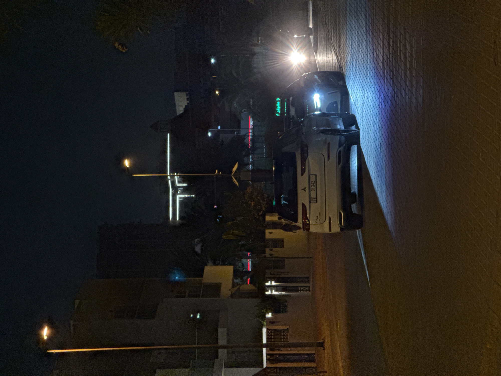 The image depicts a street scene at night in Dubai, United Arab Emirates. A white SUV, identified by a license plate with the characters "EE 31342," is parked on the right side of a paved street, with its headlights casting a bright blue glow onto the road. Another vehicle, a taxi with a green illuminated sign on its roof, is visible behind the SUV, with its headlights also illuminated. Buildings are present on the left and in the background, some of which are illuminated. A street lamp is visible on the left side of the image. The environment is dark, indicating nighttime, and the ground appears to be a patterned paving stone surface.