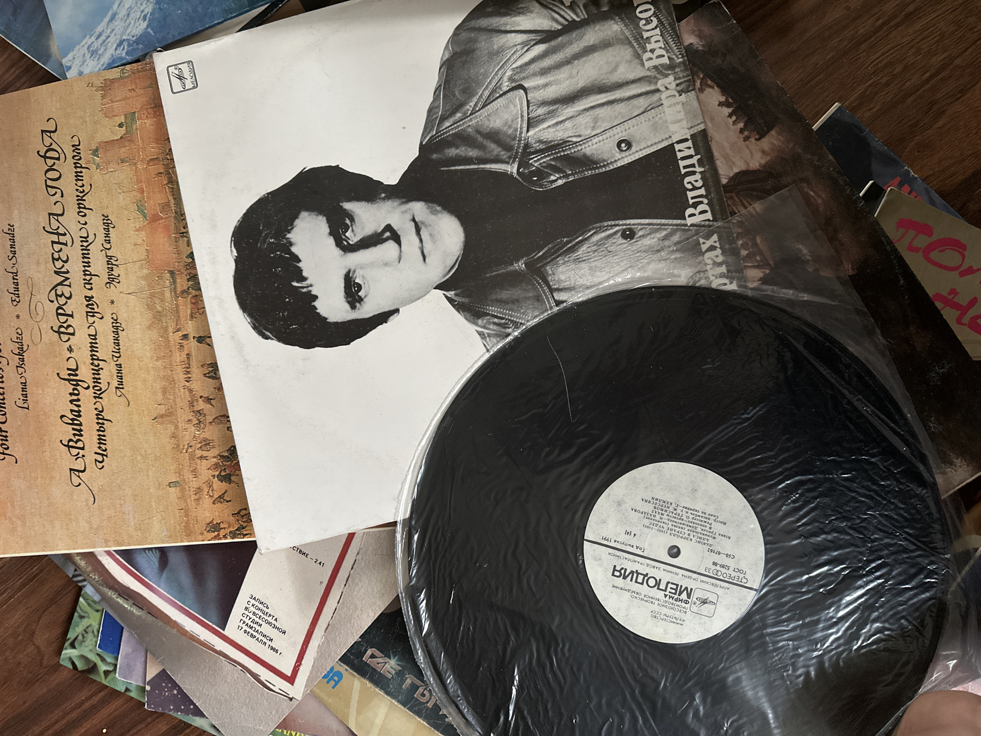 The image shows a stack of vinyl records on a wooden surface. Visible are record covers featuring musical performances: "Vivaldi: The Four Seasons" (presumably a Soviet Melodiya label pressing), a Vladimir Vysotsky album (in Russian, likely a compilation of his songs), and other records with visible Cyrillic script. A black vinyl record, still in its plastic sleeve, covers part of Vysotsky's face on his record cover. The record label on the vinyl is a Melodiya pressing. One of the vinyl record sleeves has a date "17 февраля 1986 г." (February 17, 1986), implying that it might be a recording of a concert. The year 1991 is also on the record label. The setting is not specific to Nizhny Novgorod; the records and their labels indicate Soviet or Russian origin, but the location cannot be determined from the image.