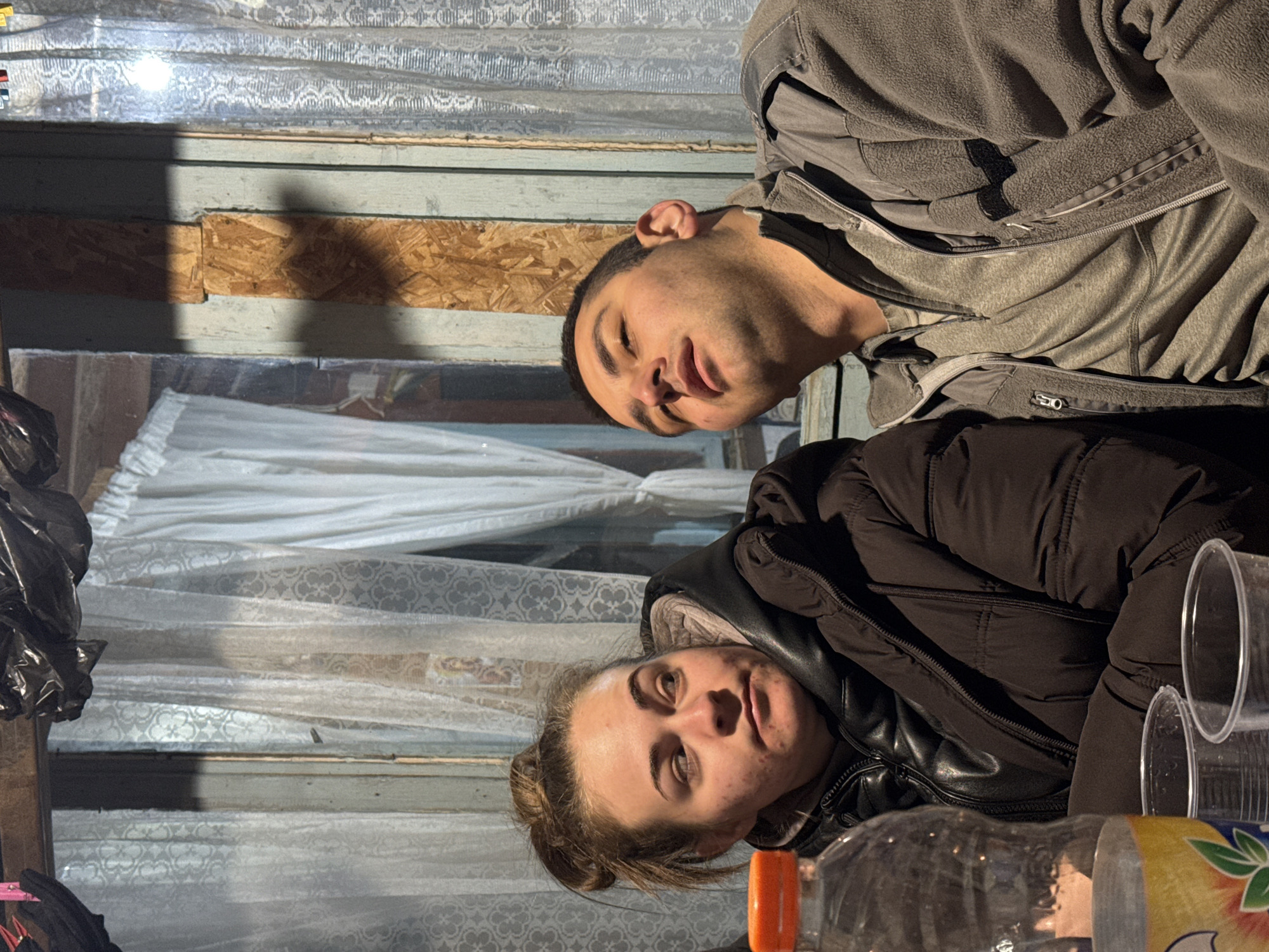 Two individuals, a male and a female, are indoors. The male is wearing a gray jacket. The female is wearing a dark jacket. The background includes a window covered with patterned curtains and a section of boarded-up wall. There are plastic cups and a bottle on a surface in the foreground. The image does not provide enough information to determine if it was taken in Kondolovo, Bulgaria.