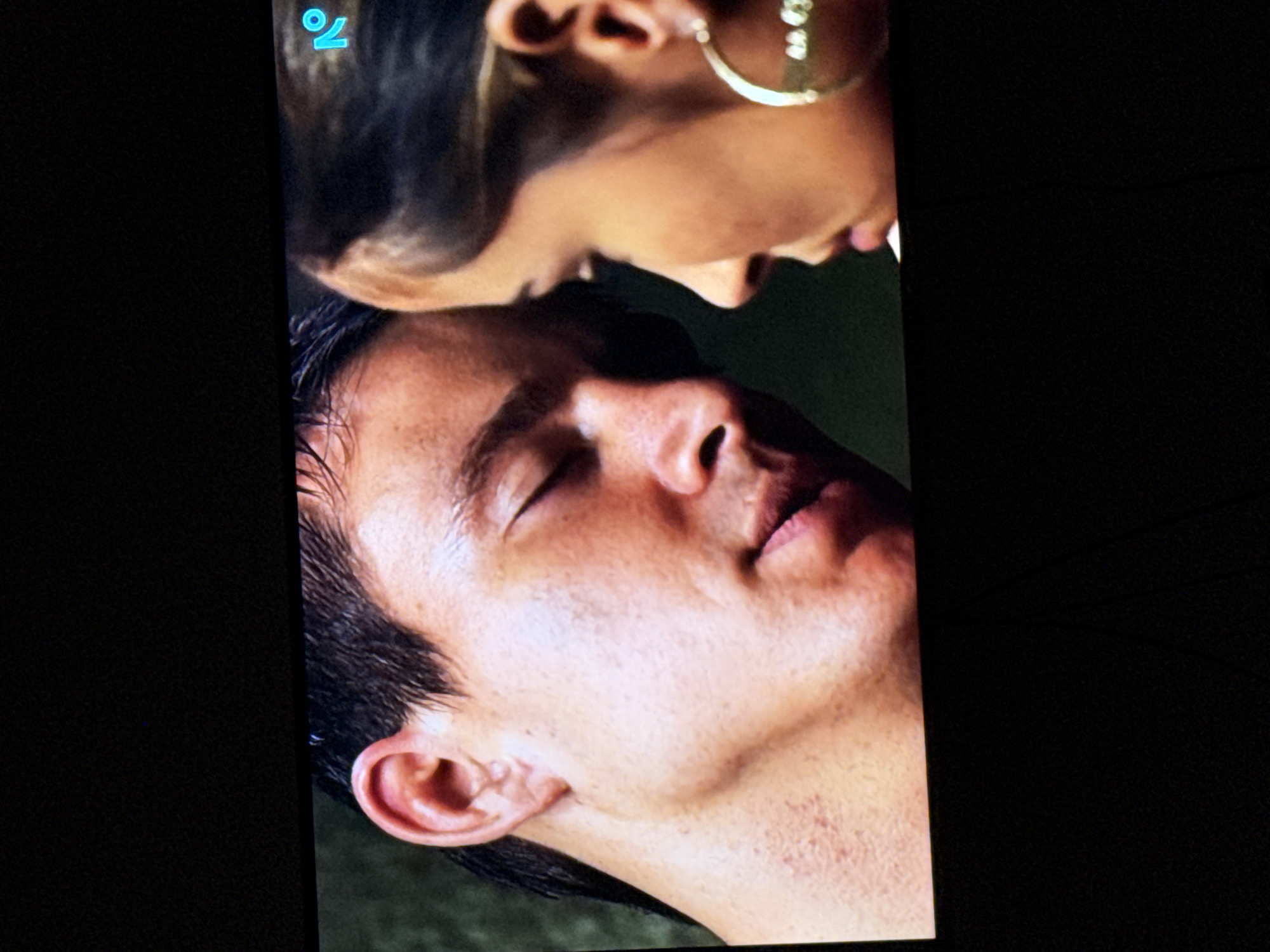 The image shows a close-up of two people, a man and a woman, with their faces near each other. The man's eyes are closed and he is on the left side of the frame. He has dark hair and appears to be sweating. The woman is on the right side of the frame and is looking at the man. She has dark hair and is wearing a large hoop earring. There is a blue logo in the upper right corner of the image. The background is dark. The image is a screen capture, likely from a movie or television show. There is no information available to link this image to Delmenhorst, Germany.
