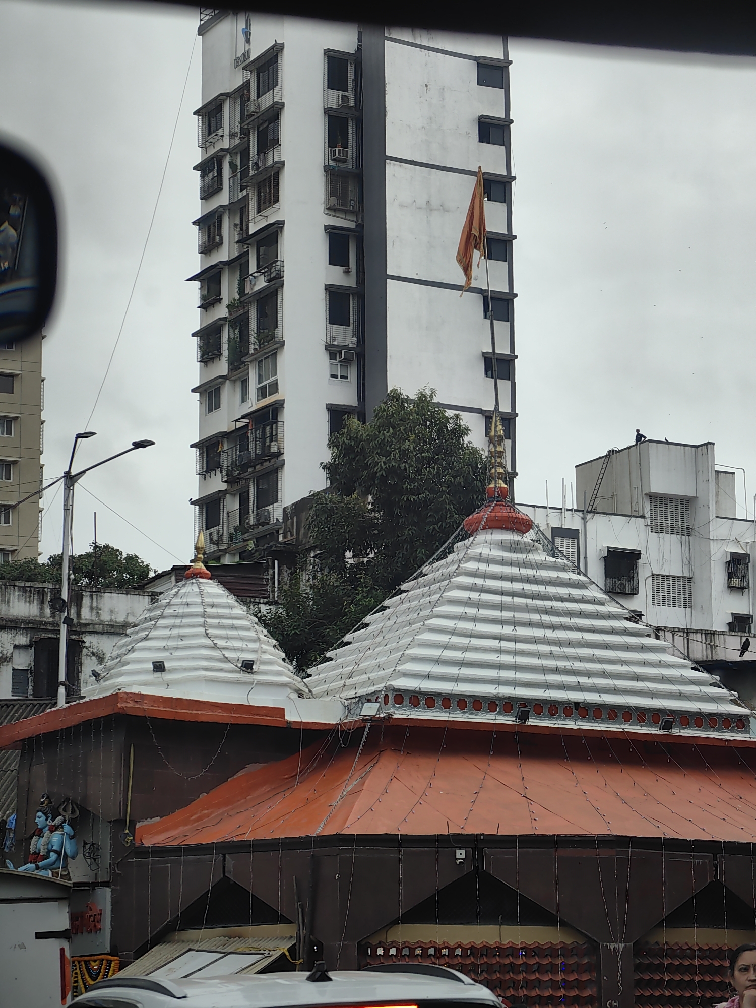 The image depicts a view of a street scene in Mumbai, India, featuring religious structures and urban architecture. In the foreground, two tiered temple domes are visible, one in the foreground and another slightly behind it. The domes are white with a red base and adorned with small lights. A statue of a blue deity, likely Lord Shiva, is visible to the left of the foreground dome. Behind the temple structures, a tall, white building dominates the skyline, characterized by numerous windows with balconies and air conditioning units. A saffron-colored flag flies from a pole attached to this building. To the left of the tall building, a street light is visible. Further to the right, another white building with multiple windows and what appear to be ventilation grills can be seen. The sky is overcast, suggesting a cloudy day. A partially visible car or vehicle is in the lower portion of the frame, obscuring the ground level details. A woman is visible in the lower right corner of the image, looking to the left. The scene suggests a typical urban environment in Mumbai with a blend of religious and residential/commercial structures.