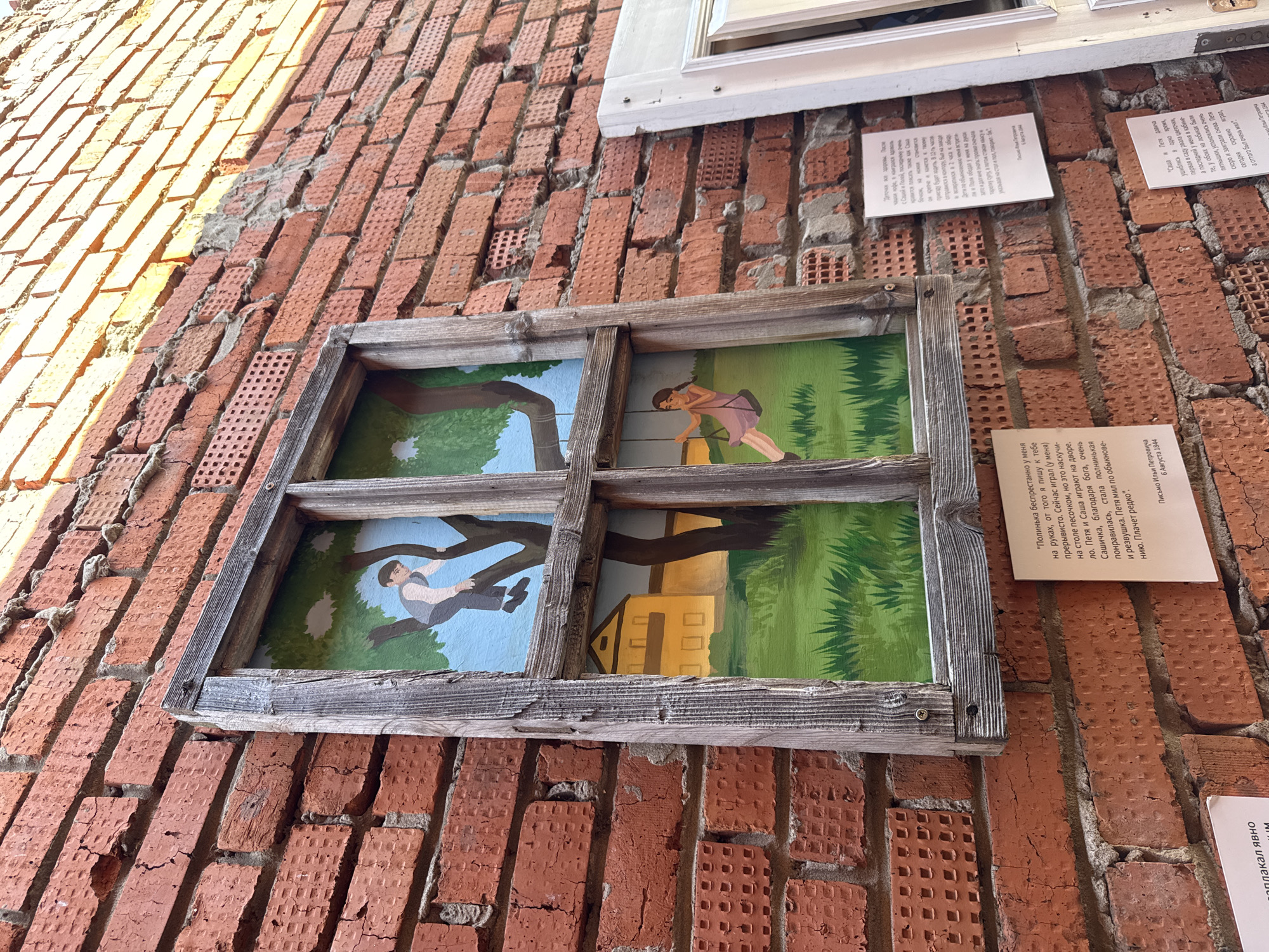 A medium, eye-level shot shows artwork mounted on a red brick wall in Votkinsk, Russia. The artwork, framed to look like a window, is divided into four panels depicting scenes of children. The panels show:
1. A child swinging from a tree branch over water.
2. A child using a pump.
3. A child falling out of a tree.
4. A house.
Several sheets of paper with text are mounted around the artwork. The red brick wall is visible in the background, showing texture and imperfections.