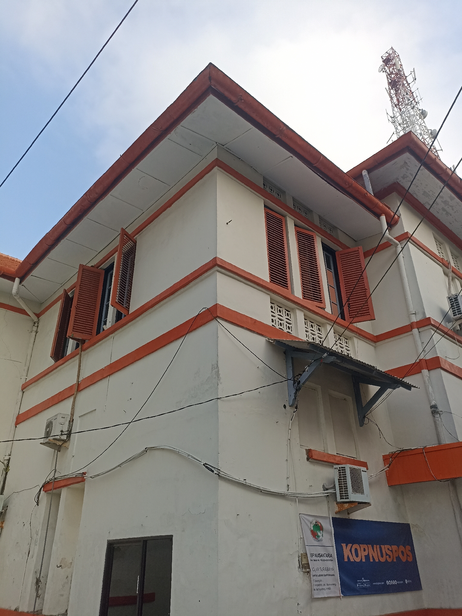 The image shows the exterior of a white building with orange trim, suggesting it is a historical or government building. The building features several windows with brown wooden shutters, some of which are open. There are also air conditioning units mounted on the exterior walls. A telecommunications tower is visible in the background, indicating an urban environment. Two banners are attached to the building's facade. One banner, with text in Indonesian, appears to be a service or information notice. The other banner, in orange and blue, displays "KOPNUSPOS". Based on the presence of "KOPNUSPOS" and the general architectural style, this building is likely a post office or a related postal service facility within Kelurahan Kepatihan, Indonesia. The sky is visible, and it appears to be daytime with some cloud cover. Electrical wires are strung across the building.
