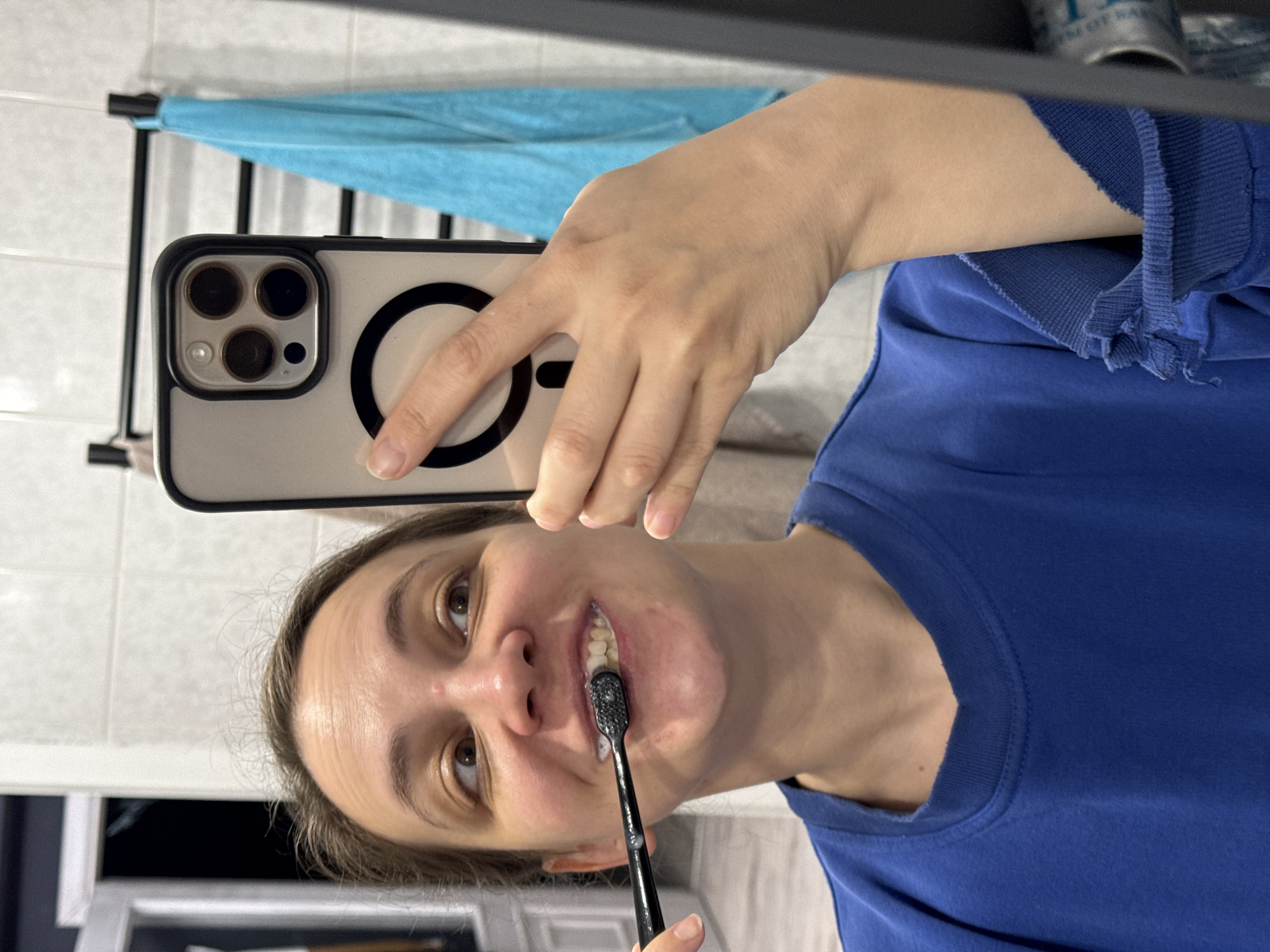 A woman is taking a selfie in a bathroom. She is holding a smartphone with a black and white case and a circular magnetic accessory. The phone has multiple camera lenses visible. She is wearing a blue t-shirt and brushing her teeth with a black toothbrush. A light blue towel is visible in the background. The location is a bathroom, though a specific location in Chaikovsky, Russia cannot be determined from the image.