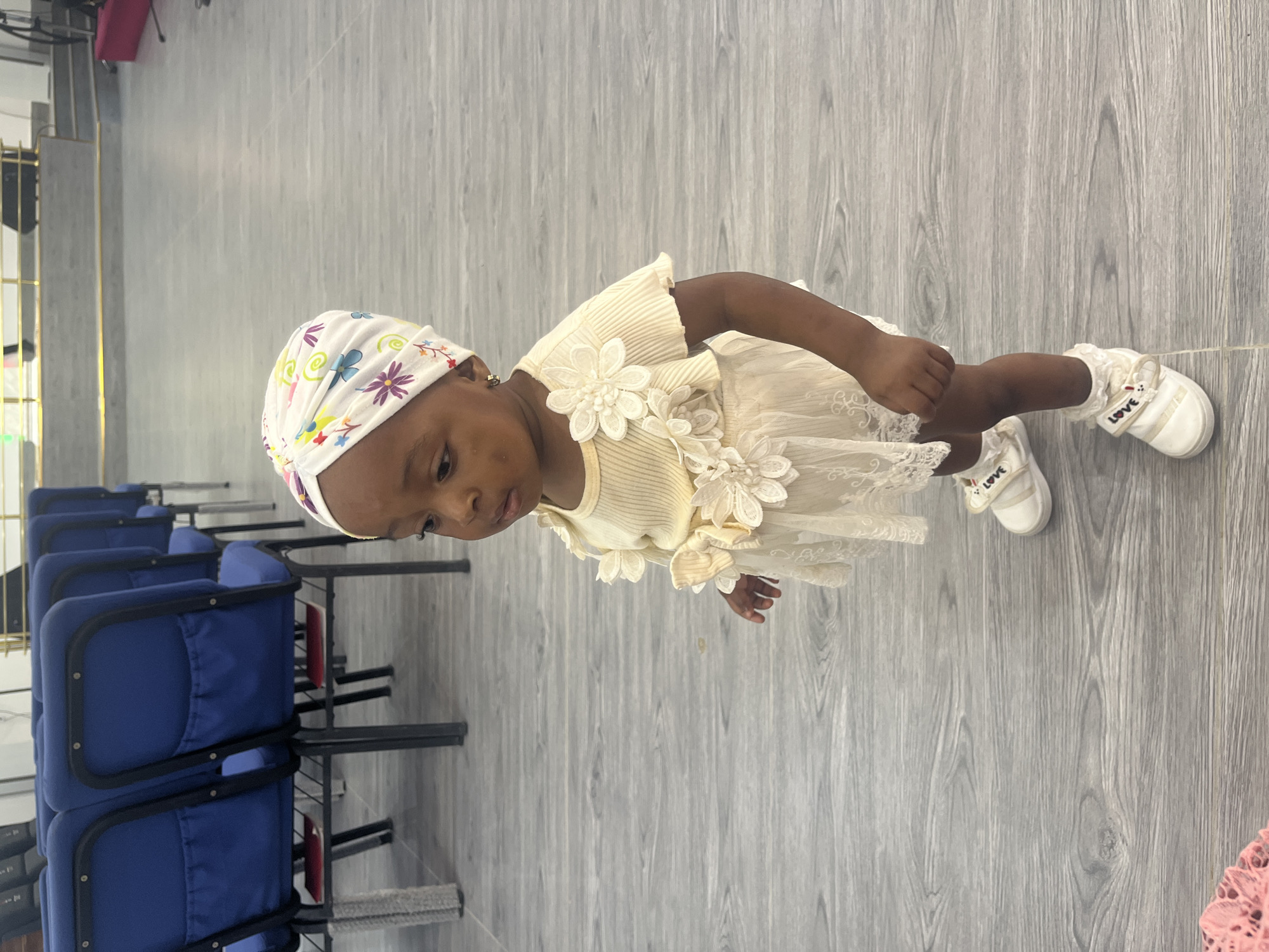 A young child stands on a grey wood-patterned floor. The child wears a cream-colored dress adorned with floral appliqués and a matching turban with a floral print. The child's feet are clad in white sneakers. In the background, rows of blue chairs with black frames are visible. The scene appears to be indoors, possibly in a church or event hall in Ado, Nigeria.