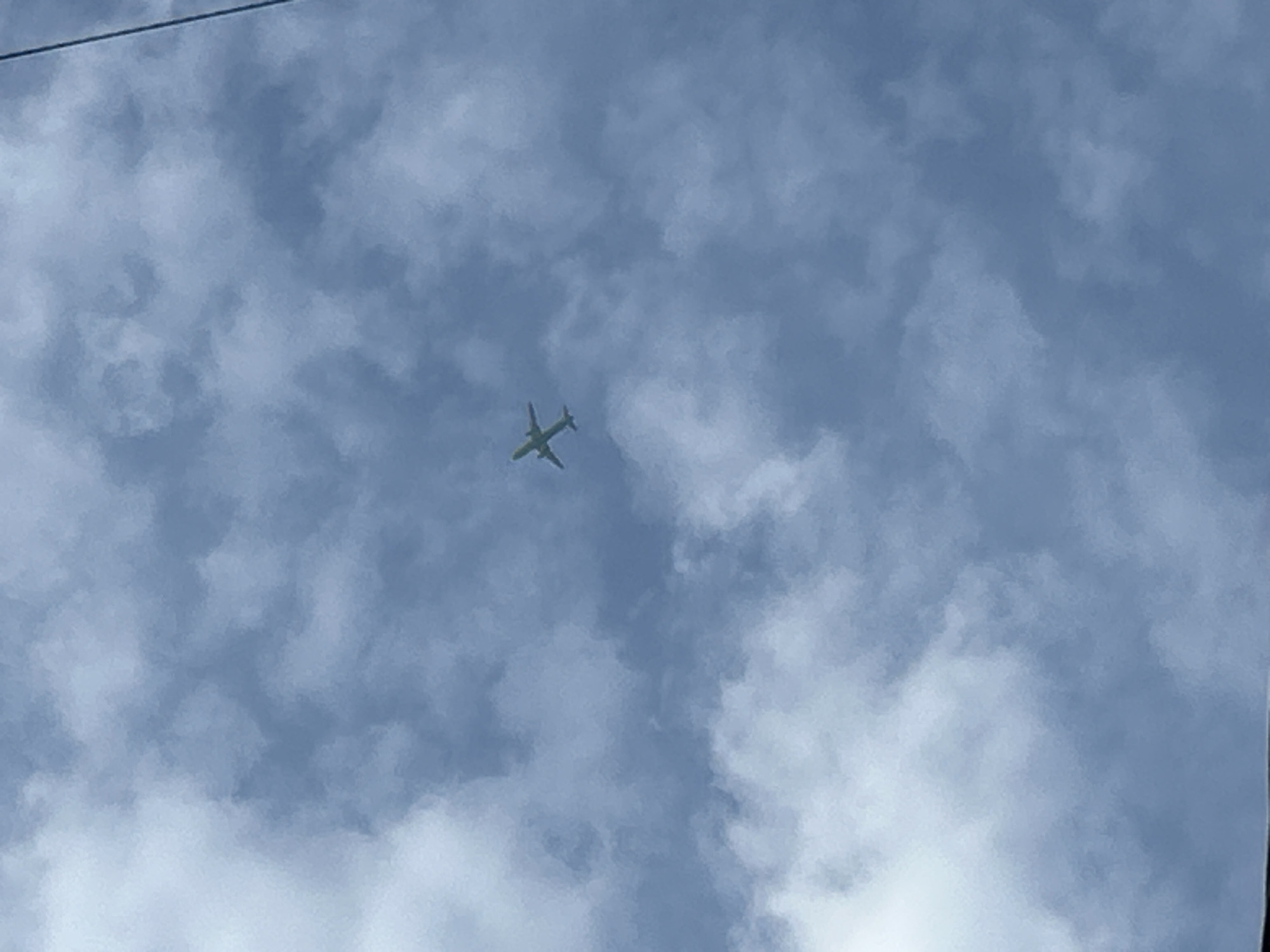An airplane is pictured from below, flying through a sky filled with clouds. The airplane is a commercial jetliner, with wings and a tail visible. The clouds are white and fluffy, with some darker gray areas. The sky is a pale blue. The image is taken from a low angle, looking up at the airplane. There are no people visible in the image.