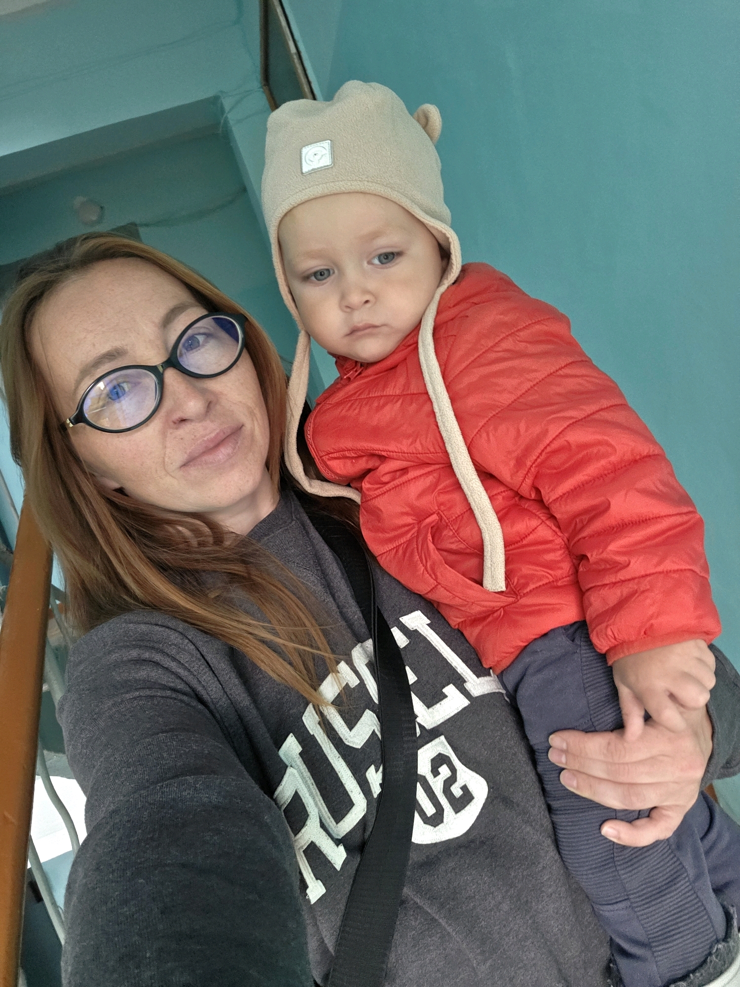 A selfie is taken by a woman with red hair and glasses, holding a baby. The woman is wearing a dark grey sweatshirt with white lettering. The baby is wearing a red puffer jacket and a light beige hat with ear details. They are standing in what appears to be a stairwell, with a turquoise wall and a wooden railing visible. The background suggests an indoor setting, possibly a residential building in the city of Chaikovsky, Russia, given the context. The lighting is natural, and the focus is on the woman and child.
