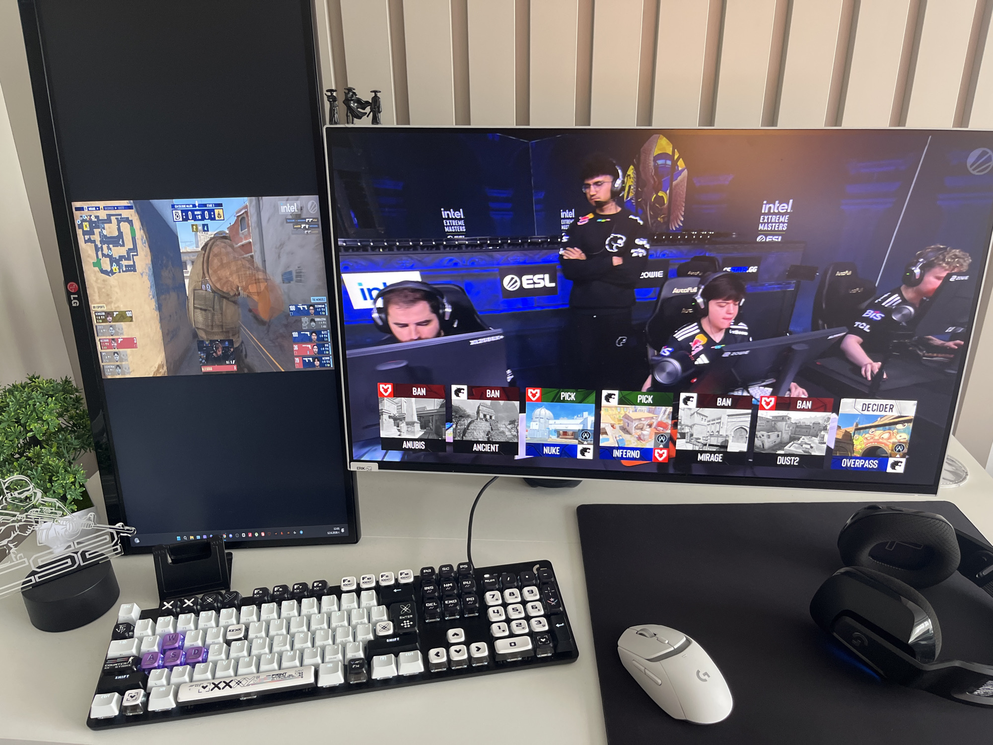 The image depicts a computer setup, likely for gaming, featuring two monitors, a keyboard, two mice, and headphones. The left monitor displays a map and gameplay from Counter-Strike, with player information and a timer. The right monitor shows a live esports broadcast, with players wearing headphones and jerseys, and a game map selection screen. The scene is set on a white desk with a black mousepad. A small decorative plant is visible on the left. The environment appears to be indoors, with a textured, light-colored wall in the background. The image is taken from a slightly elevated perspective, focusing on the computer equipment. There is no discernible information or context within the image that places it in the city of Varna, Bulgaria.