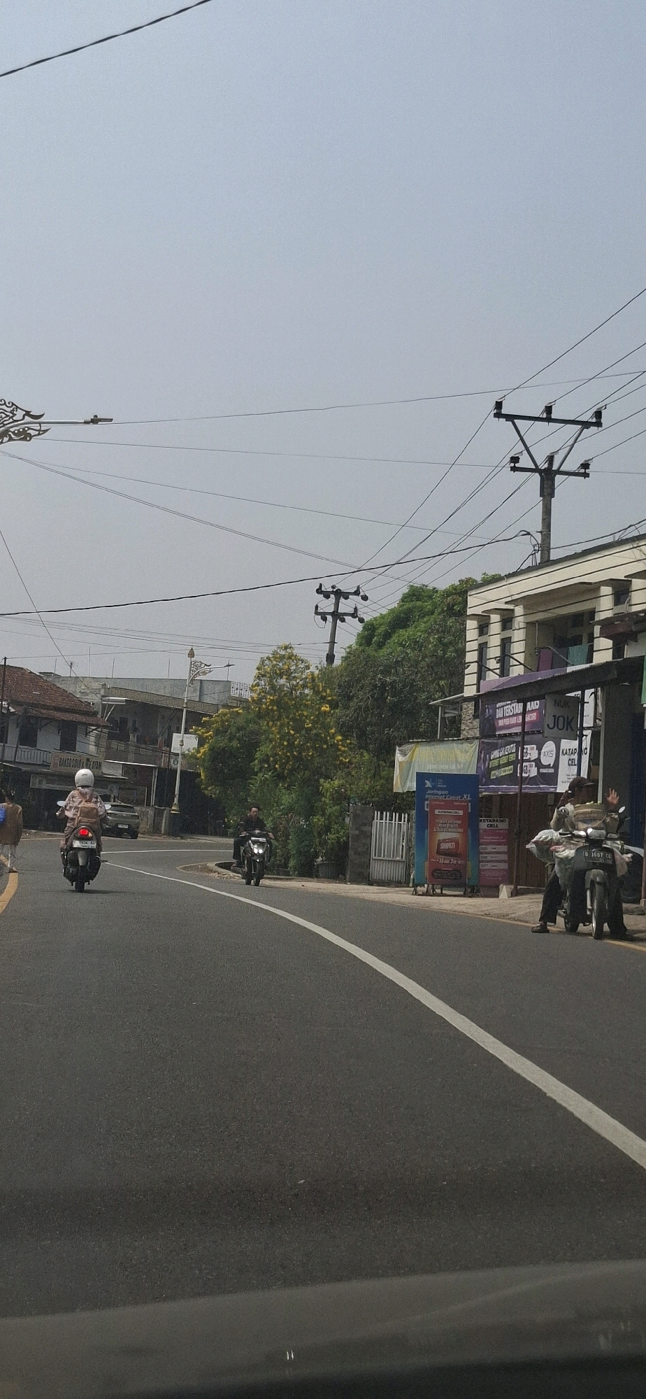 The image depicts a street scene in Susukan, Indonesia, on an overcast day. A paved road with a white lane marker curves through the frame, with buildings lining one side and a dense canopy of trees on the other. Several motorcycles are present. In the foreground, a motorcycle rider wearing a helmet and a backpack is visible from behind, traveling away from the camera. Further down the road, another motorcycle rider is moving towards the camera. On the right side of the road, near a building with a white facade and multiple pillars, two people are seated on a motorcycle. One individual appears to be waving. Several utility poles with power lines are prominent in the upper portion of the image. Advertising banners are affixed to the building. The overall atmosphere is that of an ordinary day in a semi-urban environment.