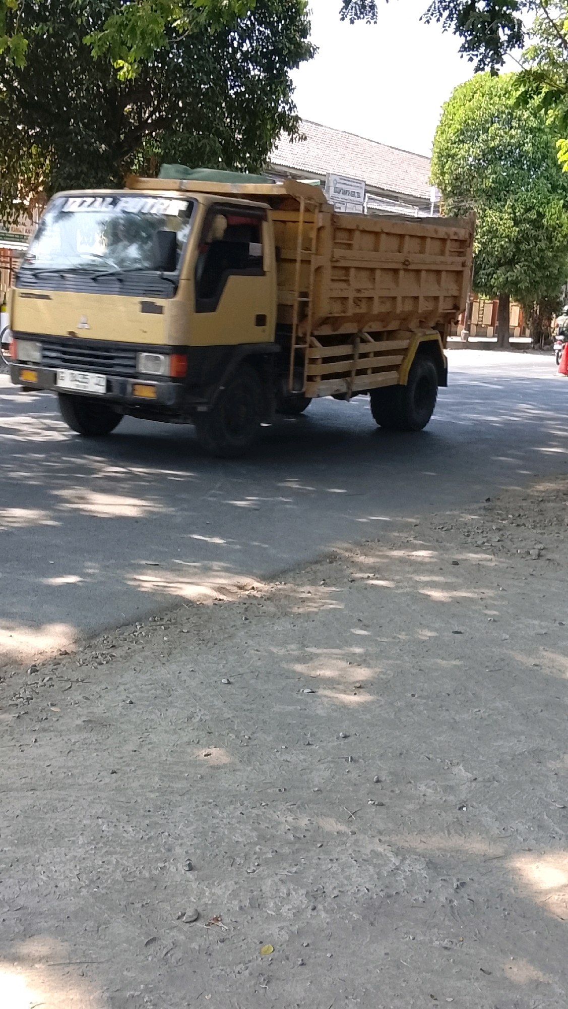 A yellow dump truck with a wooden bed is parked on a paved road. The truck has a white license plate that reads "E 8435 IZ". The scene appears to be in Slawi, Indonesia, indicated by the license plate format. The truck is parked on the right side of the road, and the environment suggests a sunny day with shadows cast by trees onto the road surface. Buildings and trees are visible in the background.