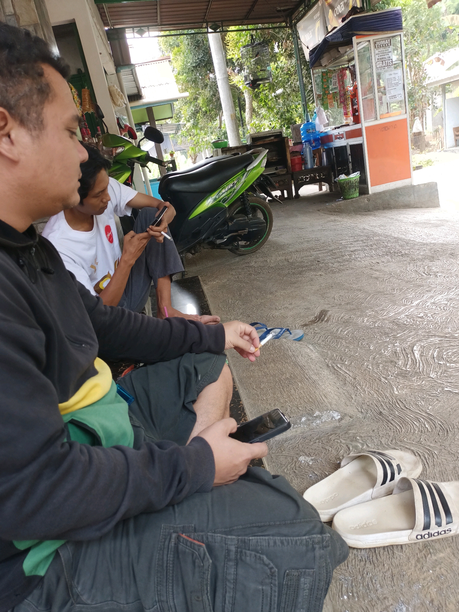 This is a medium shot, eye-level, outdoor photograph taken in the city of Warungpring, Indonesia. In the foreground, a man in a black hoodie and grey pants is sitting on a concrete floor, holding a smartphone. To his left, another man in a white t-shirt and grey pants is also sitting and holding a smartphone, with a cigarette between his fingers. Behind them, a green and black motorcycle is parked. Further in the background, there is a small food stall with orange and white accents, displaying various items. Trees and foliage are visible, suggesting a natural, slightly rural setting within the city. The overall scene depicts a casual moment of people resting and using their phones in an outdoor, public space.
