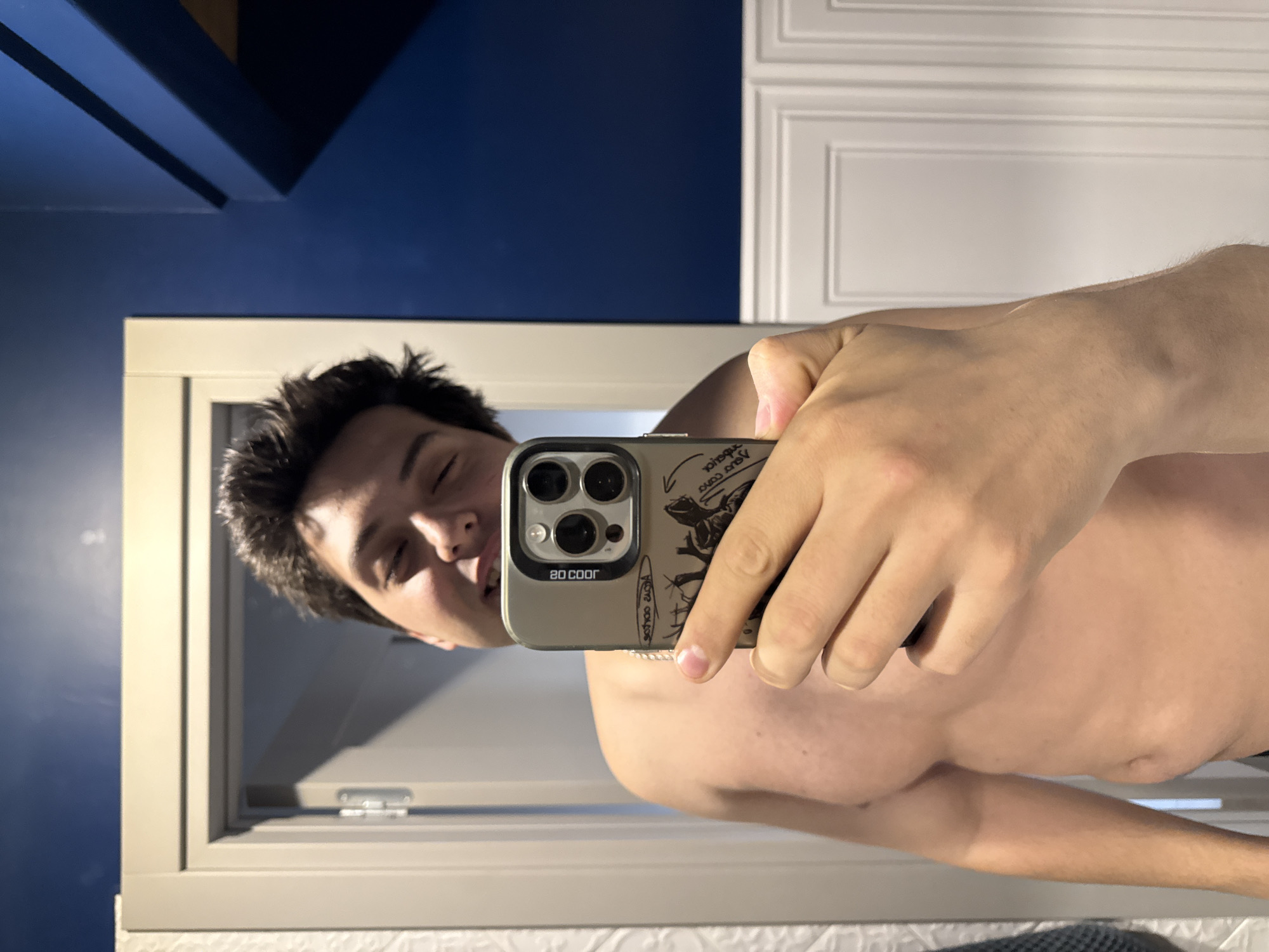 A shirtless man takes a selfie with his phone, using the reflection of a bathroom mirror. The phone has a light brown case with cartoon drawings. The bathroom has white cabinets and dark blue walls. The location is not apparent. It is not possible to determine if this is located in the city of Vidnoye, Russia.