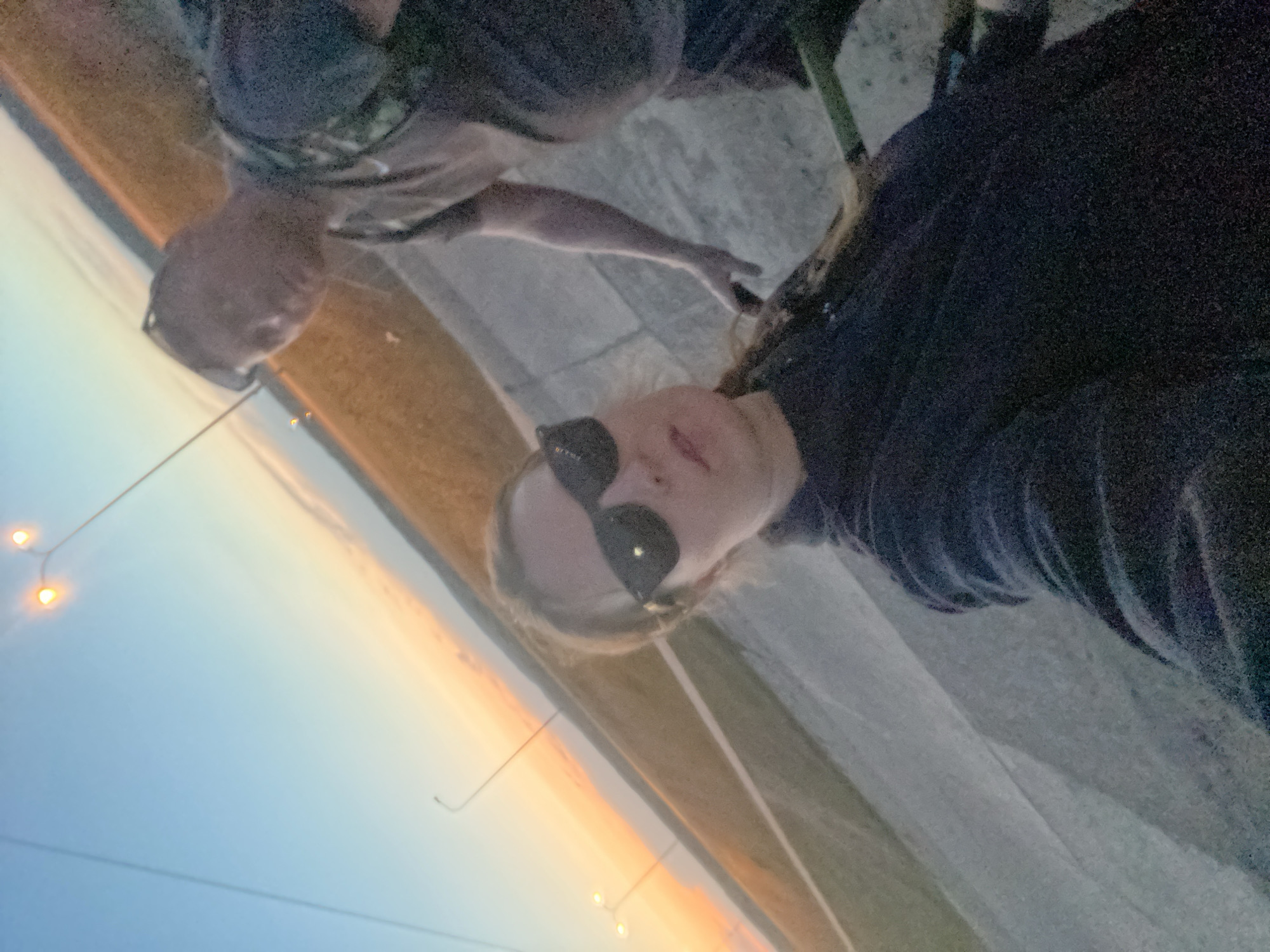 A rotated medium shot shows two individuals outside at dusk. The person in the foreground is wearing sunglasses and a dark-colored top. The other person, positioned behind and slightly to the left, is also dressed in dark clothing and wearing glasses. In the background, there is a concrete surface and a sky exhibiting colors of dusk. Hanging lights are visible. The location is suggestive of an outdoor setting, possibly in Chaikovsky, Russia.
