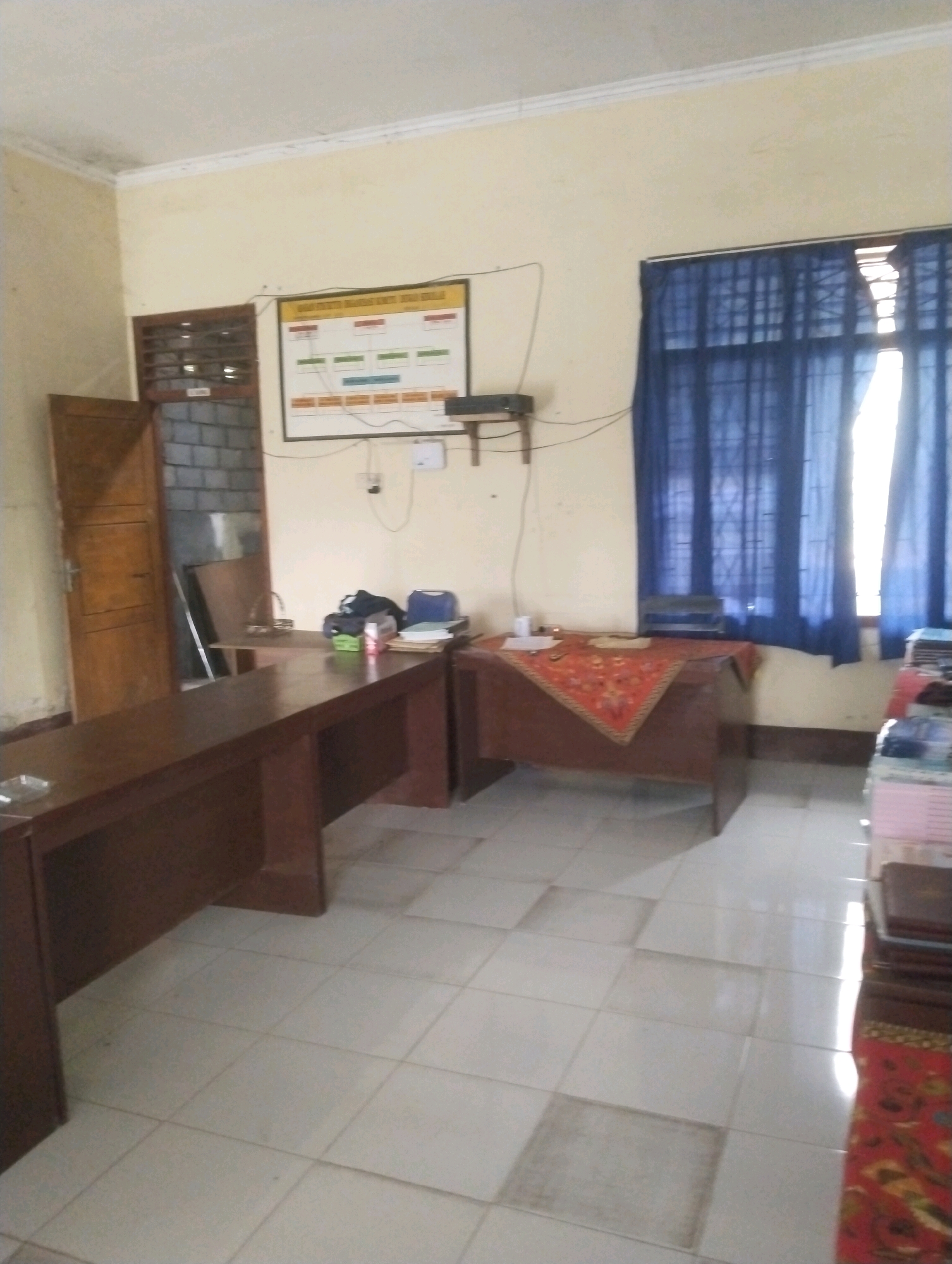 This is a medium-shot, eye-level, indoor photograph of an office space. The room has cream-colored walls and a tiled floor. In the foreground, to the left, is a long, dark brown wooden desk with a rectangular hole in the center. To the right of this desk is another, smaller, dark brown wooden desk covered with a red and gold patterned cloth. On this desk, a stack of papers and a small white object are visible. Behind this desk, a blue curtain is drawn across a window. In the background, to the left, is a wooden door ajar, revealing a brick wall and some shelving. A framed organizational chart is mounted on the wall above the desks. A small black electronic device is attached to the wall below the chart. The overall impression is of a functional, if somewhat dated, office in Kareng Baru, Indonesia.