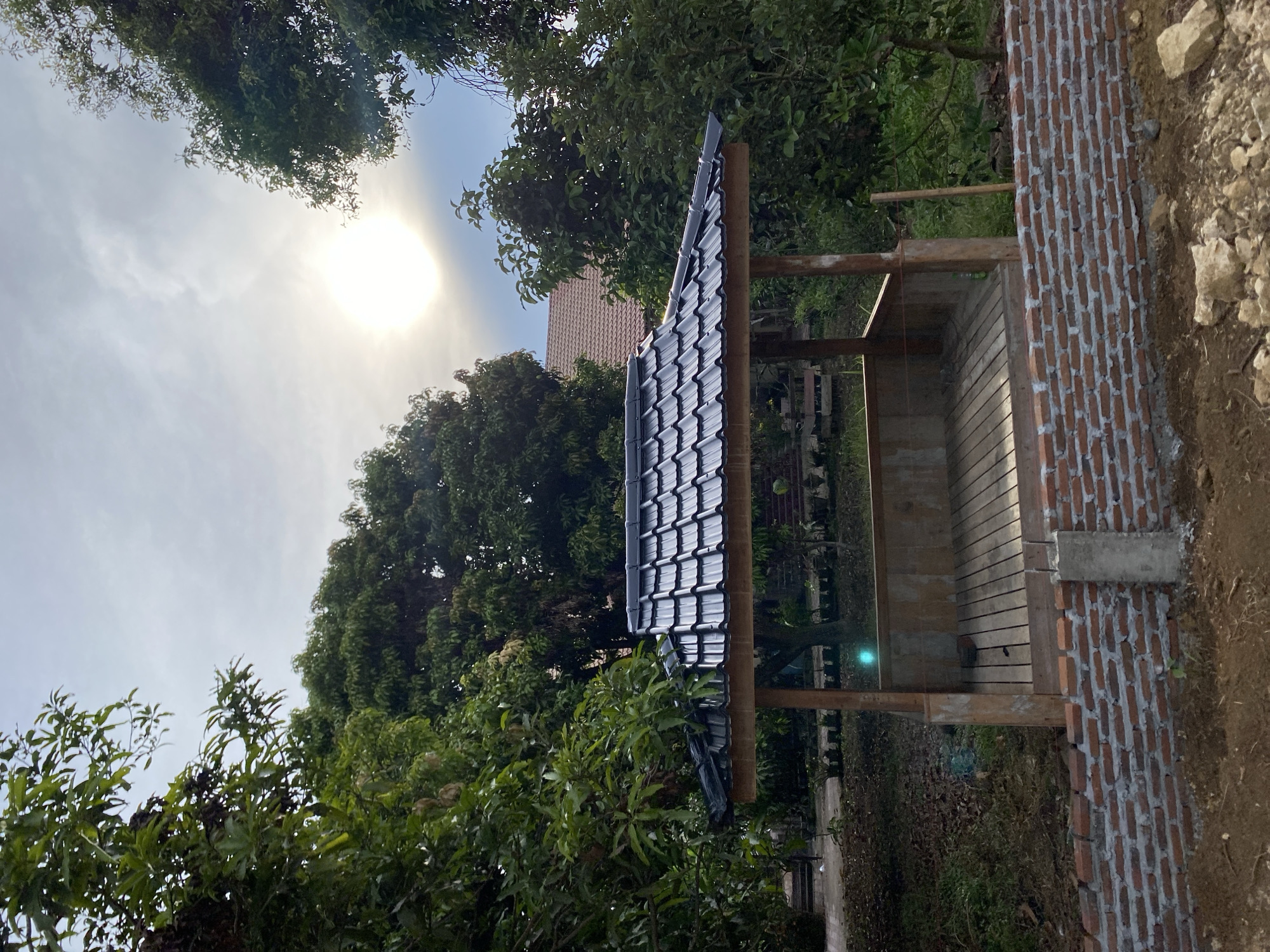 This image captures an outdoor scene under a bright, hazy sky, likely during daytime. The sun is a prominent, diffused orb in the upper left quadrant, suggesting a moderately sunny day with some atmospheric diffusion. The setting appears to be a semi-developed outdoor area, possibly a garden or construction site, with lush green foliage from trees and plants dominating the left and background.
The main subject in the foreground is a newly constructed, open-sided structure. It features a dark-tiled roof supported by wooden beams. The floor of this structure is made of horizontal wooden planks. To the right of this structure, there is a partially built brick wall, constructed with reddish-brown bricks and light gray mortar. The wall appears to be a boundary or part of a larger construction. The ground around the structure is a mix of dirt, stones, and some dry leaves, indicating an outdoor environment that is either under development or in a natural state.
A building with a reddish-tiled roof is visible in the mid-ground, behind the new structure and partially obscured by trees. The overall impression is one of an outdoor space being prepared or enhanced, with construction elements alongside natural vegetation. The lighting and bright sky suggest it is either morning or late afternoon. There are no people visible in the image. No visible text is present.