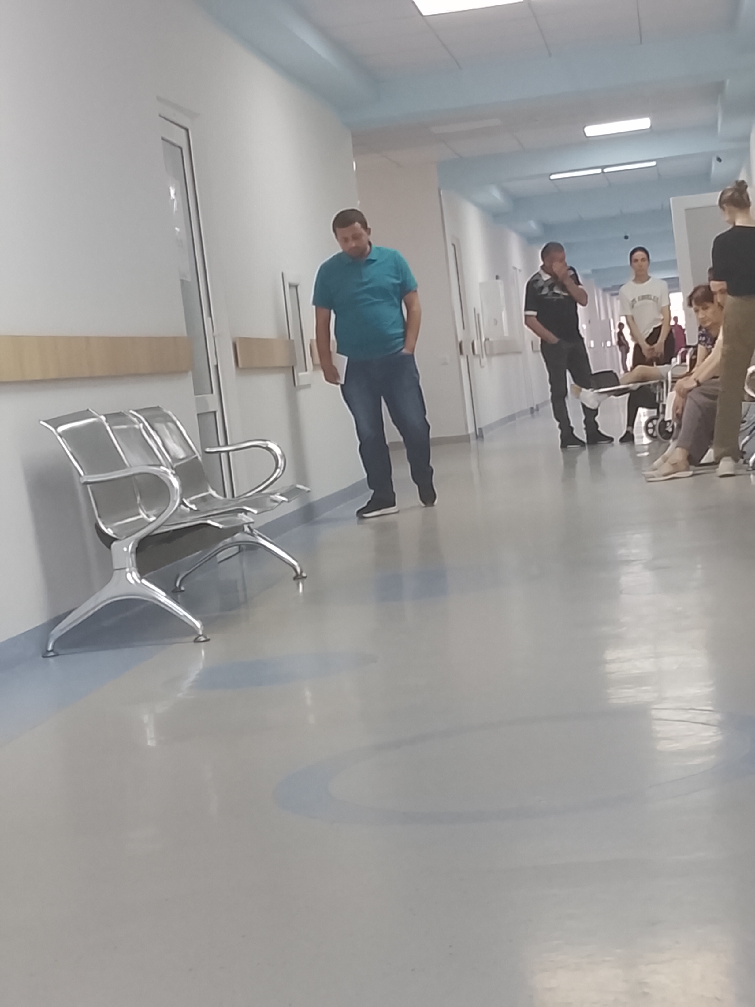 The image depicts a hospital corridor. A man in a teal polo shirt and jeans stands in the foreground, looking down. To his right, a group of people are gathered. A man in a black t-shirt and jeans stands speaking on his phone. A woman in a white t-shirt and jeans stands next to him. Further down the corridor, a woman in a brown top and patterned skirt is seated, and a woman in a white t-shirt and dark pants is partially visible. To the left, a silver, modern waiting bench is positioned against a white wall. The corridor features a polished, light-colored floor with subtle blue circular markings. Fluorescent lights are visible on the ceiling. The overall environment appears to be a clean, well-lit medical facility.
