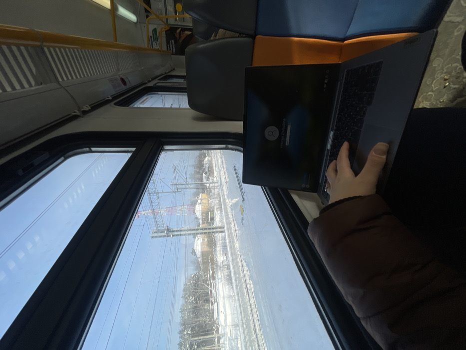 An interior view from a train carriage depicts a snowy landscape outside the window, located near Fryazevo, Russia. A person, whose right arm in a brown jacket sleeve and hand with a ring are visible, operates a dark grey laptop resting on their lap. The laptop screen displays a login prompt featuring a user profile icon and the name "Victoria Zhulyabina." Outside the window, a clear blue sky spans over snow-covered ground and railroad tracks. In the midground, several light-colored buildings are visible among snow-dusted trees, with a distinctive red and white lattice telecommunications tower in the distance. Overhead power lines for the electrified railway cross the frame. Inside the train, light-colored walls, ribbed ceiling panels, and portions of grey and orange/blue upholstered seats are discernible, along with yellow grab handles and overhead lighting.