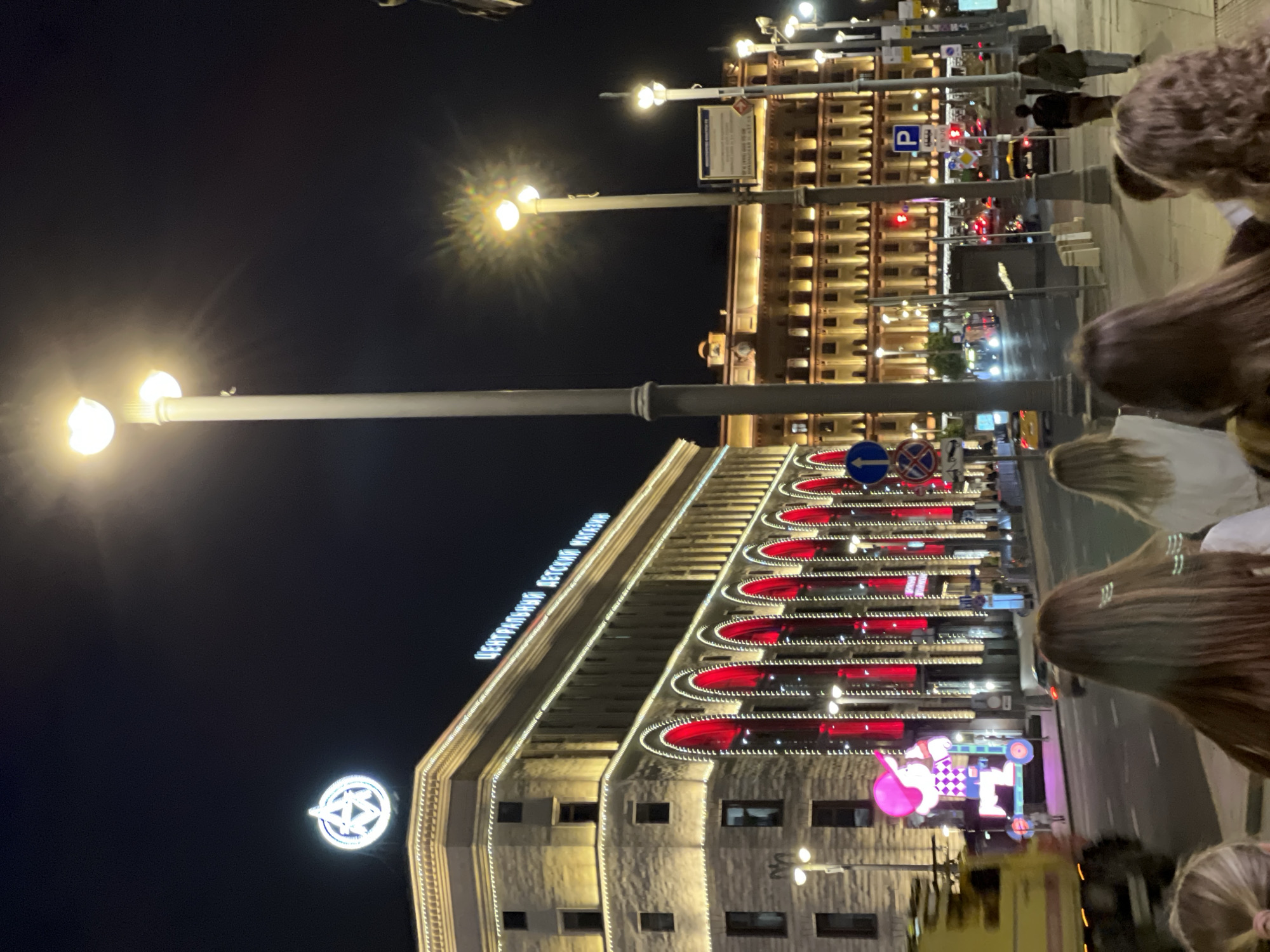 The image shows a night scene on a city street in Moscow, Russia. In the foreground, the backs of several people's heads are visible as they walk away from the camera. Behind them, a large building with illuminated arches and decorative lights dominates the scene. A sign with Cyrillic text is visible on the building. Further down the street, more illuminated buildings and streetlights are present. Traffic, including cars and what appears to be a construction vehicle, is visible on the street. The overall atmosphere is that of a busy urban environment at night. The architecture suggests a historical or grand building, possibly a prominent landmark or commercial establishment. The lighting is predominantly artificial, with streetlights, building lights, and vehicle headlights contributing to the illuminated scene.