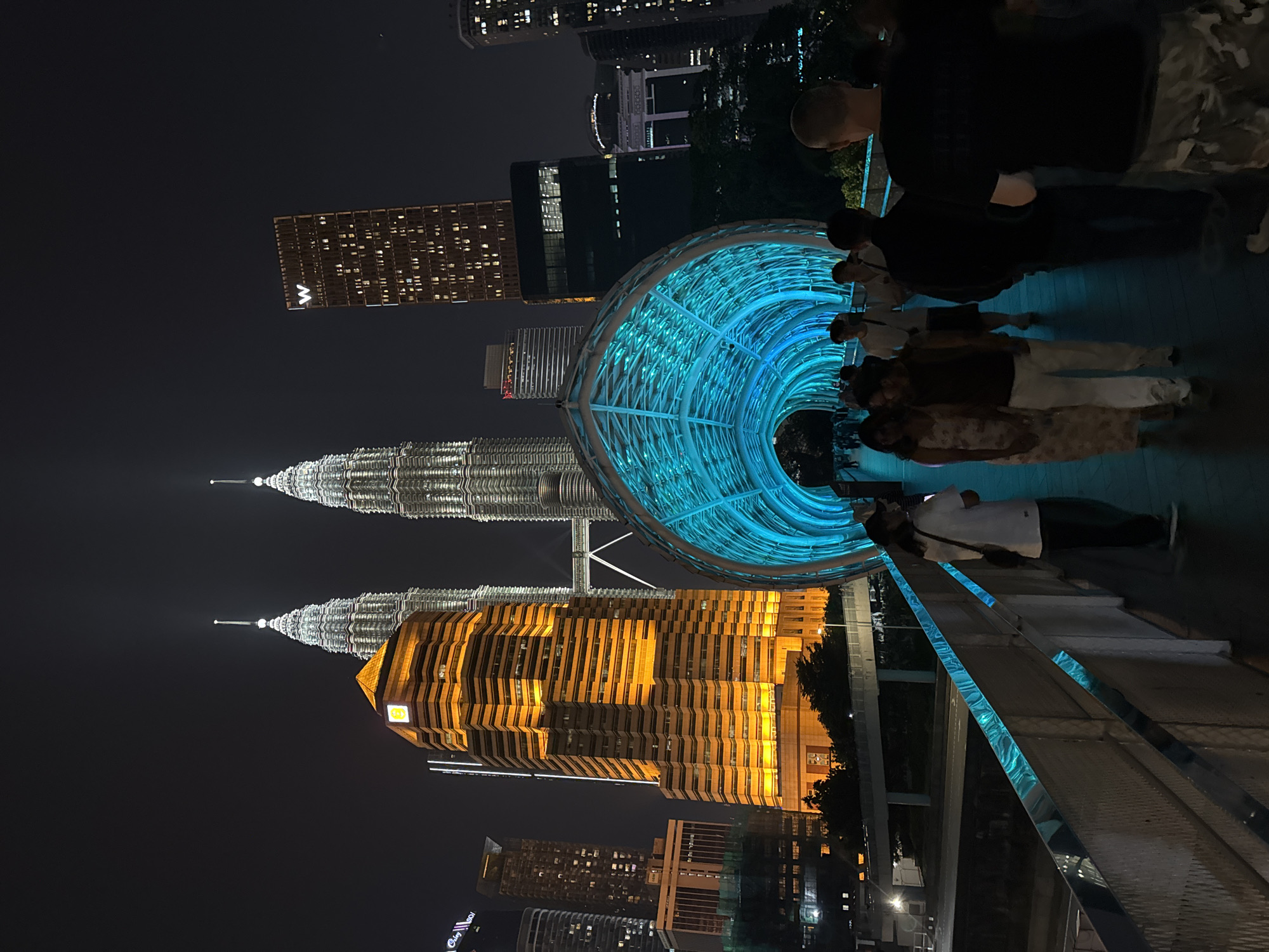 The image shows a nighttime view in Kuala Lumpur, Malaysia, featuring the Petronas Twin Towers illuminated in orange and silver. A blue-lit pedestrian bridge or walkway is visible in the foreground, with several people walking through it. The surrounding cityscape is dark, with other buildings illuminated in the distance. The bridge's design is modern and tubular, creating a visual tunnel. The scene appears to be a popular tourist spot, with people pausing to take in the view of the skyline.