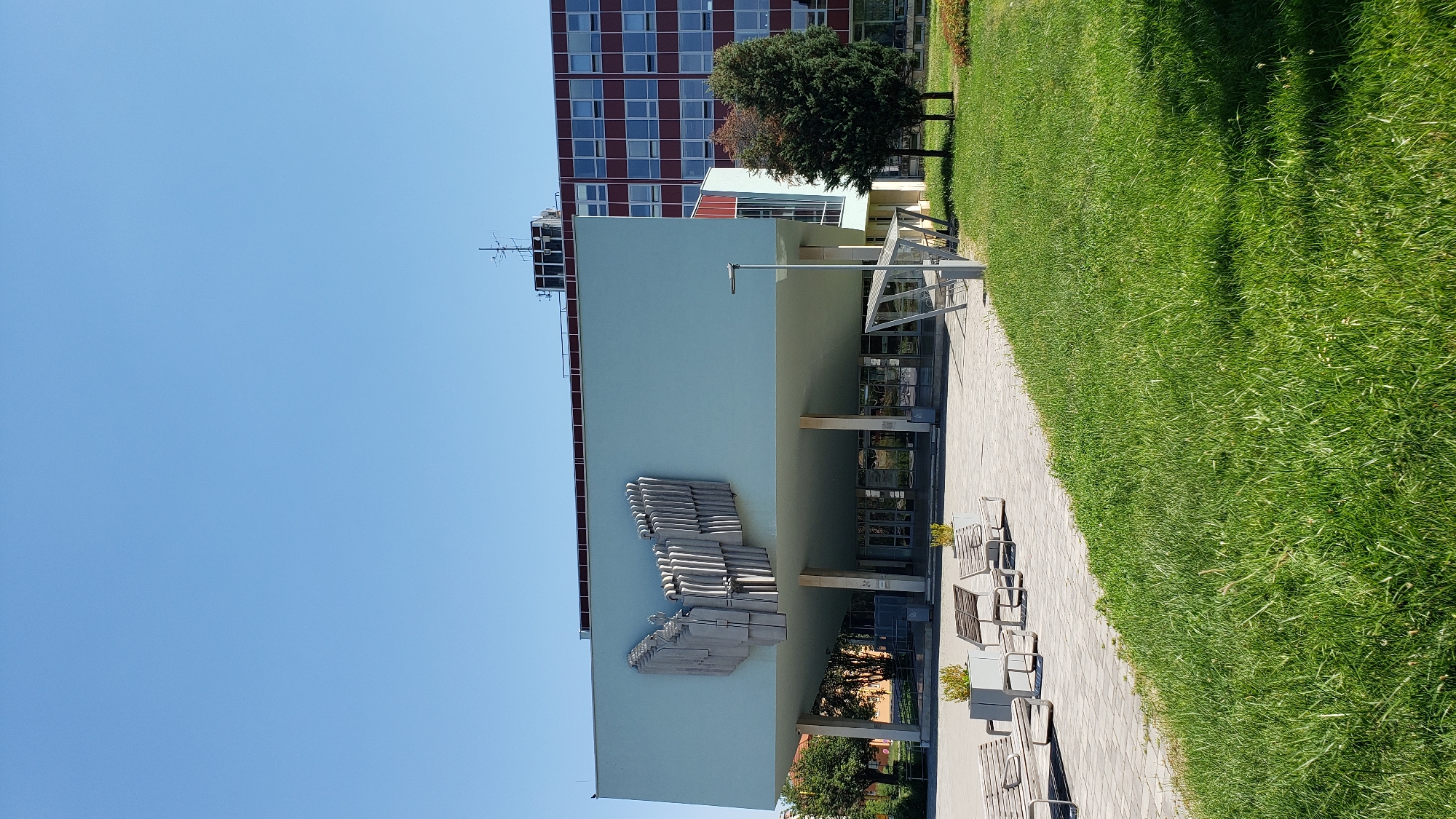 The image depicts the exterior of a modern building with a light blue facade, featuring a prominent relief sculpture on its wall. The sculpture appears to be abstract, composed of interlocking geometric shapes. To the left of the building, a taller, older building with a reddish-brown exterior and a grid of windows is visible. A well-maintained lawn with green grass stretches along the right side of the frame, bordering a paved walkway. Several park benches are arranged along the walkway, some with small potted plants.
In the background, trees are present, including a mature evergreen and a deciduous tree with some autumnal color. The sky is clear and blue, suggesting a sunny day. The lighting indicates it is daytime, likely late morning or early afternoon, given the shadows cast on the grass. The setting appears to be an urban or campus environment, possibly near an educational institution or a public facility, given the architecture and the presence of outdoor seating. No people are visible in the scene. The image is oriented vertically, with the sky at the left and the ground at the right.