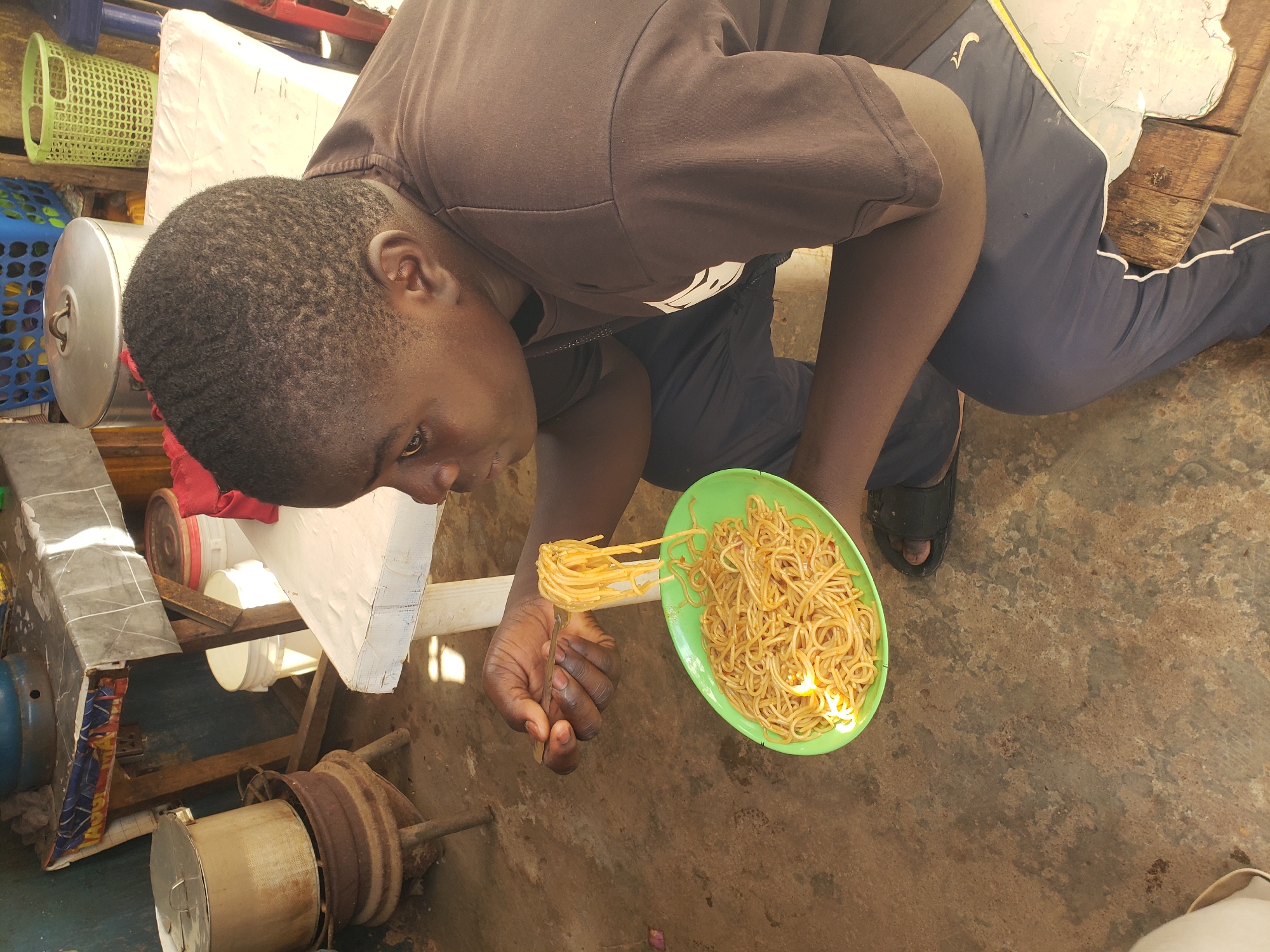 In Jos, Nigeria, a young Black individual, facing right, is seated on a wooden bench, eating a plate of spaghetti with a fork. The spaghetti is piled high on a green oval plate, with a forkful being lifted towards the individual's mouth. The person is wearing a dark brown t-shirt and dark blue sweatpants. The environment appears to be an outdoor setting, possibly a market or roadside eatery, with concrete flooring. In the background, there are various objects, including a large metal pot on a charcoal stove, a white table with a patterned cloth, and plastic baskets of different colors. The scene is lit by natural sunlight.