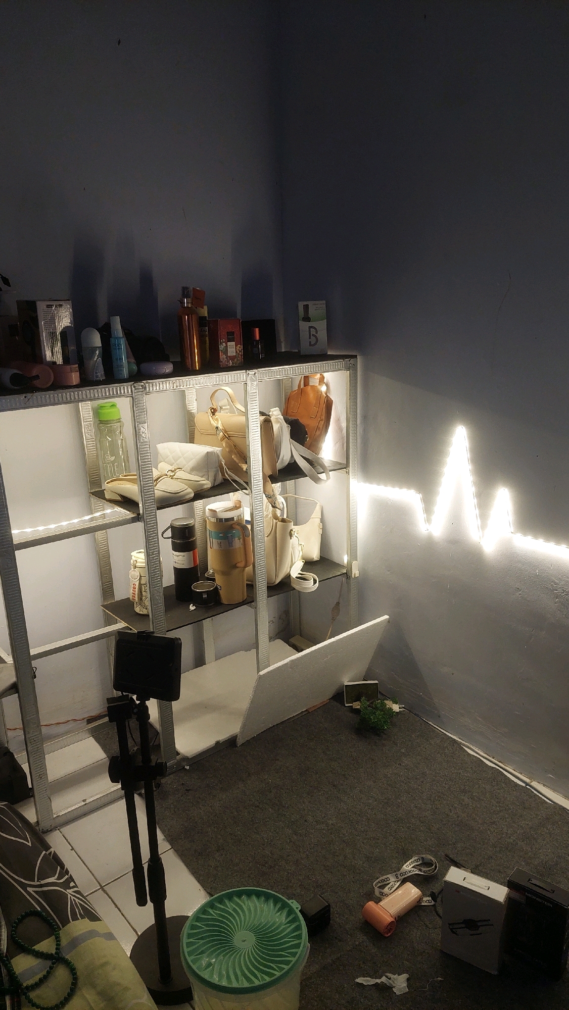 The image depicts an indoor scene, likely a room or storage area, featuring a metal shelving unit against a gray wall. The shelving unit is illuminated by white LED strip lights, which also form a heartbeat-like pattern on the adjacent wall.
On the top shelf of the unit, several items are visible, including bottles of cosmetics or personal care products, and a box with the letter "B" on it. The shelves below contain various bags in neutral tones, a water bottle, thermal mugs, and smaller containers. A black tripod with a smartphone mount is positioned in front of the shelving unit.
The floor is covered with a dark gray carpet, upon which several objects are scattered, including a green plastic container, a pink object that appears to be a portable charger or similar electronic device, and two boxes. A small artificial plant is placed in the corner near the wall. The overall lighting is dim, with the LED lights providing the primary illumination. The scene is devoid of any people. The image is presented as if it were taken within the city of Babadan, Indonesia, though there are no visual cues within the image itself to confirm this specific location.