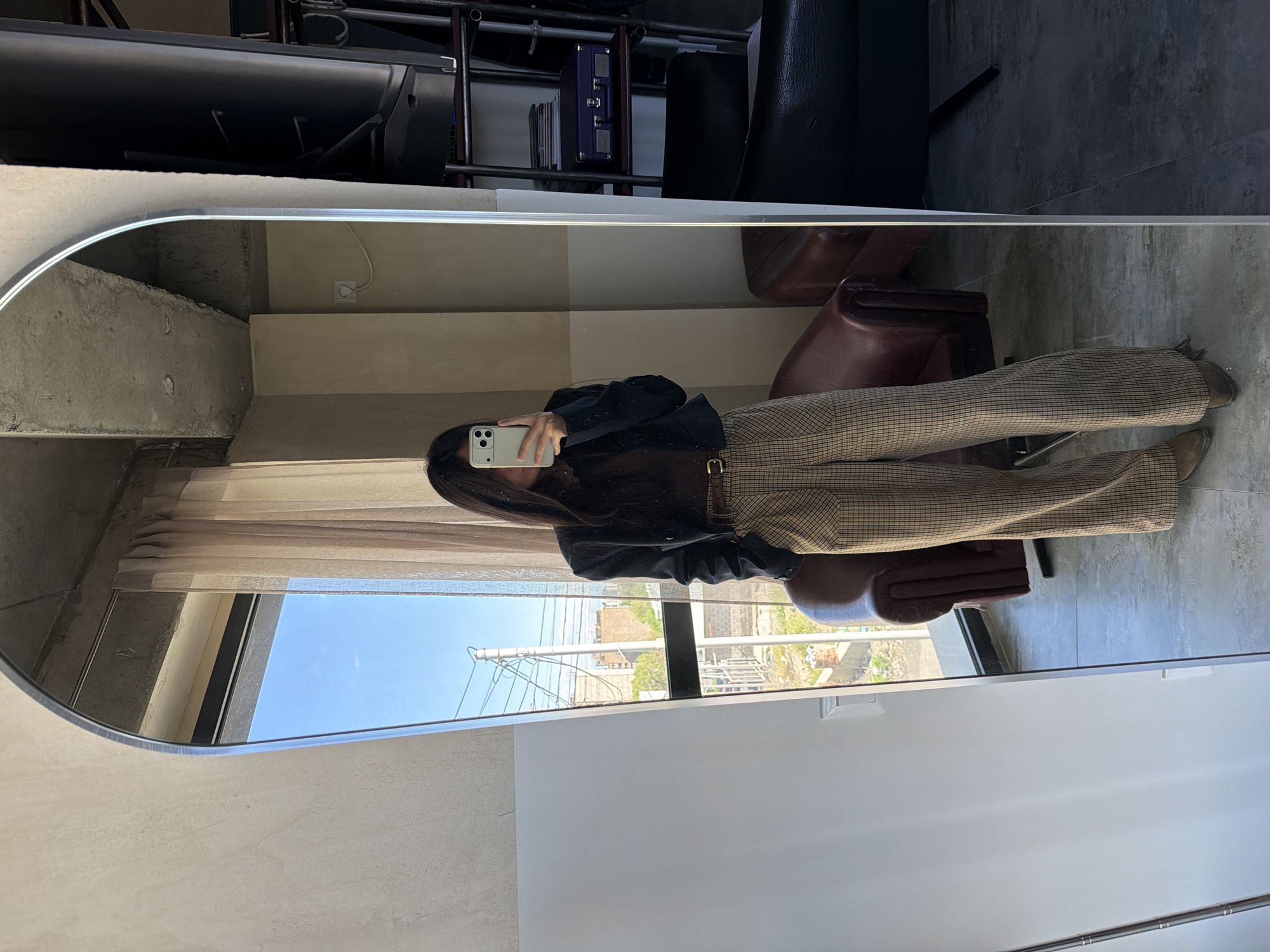 The image shows a reflection in a mirror of a person taking a selfie. The person is wearing a black top, checkered pants with a brown belt, and brown shoes. They are holding a white smartphone in their hand. The mirror reflects the interior of a room with a window showing a cityscape and a blue sky. Outside the window are various buildings, power lines, and what appears to be construction. The person is standing on a light grey tile floor, in front of a brown leather chair. There is a white wall to the right, and a textured grey wall to the left. The location is Yerevan, Armenia.