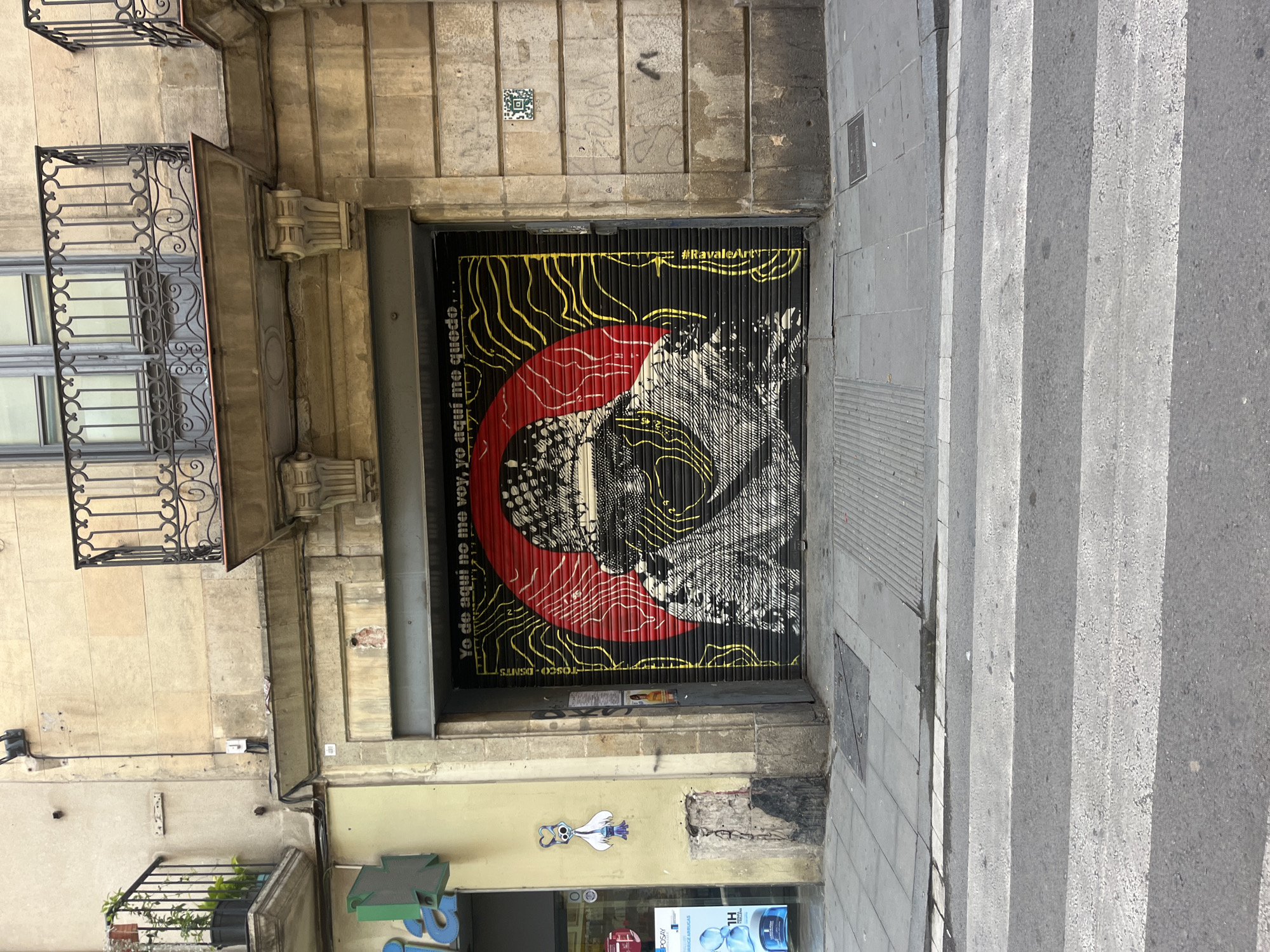 The image depicts a street view in Barcelona, Spain. The focus is on a closed storefront entrance with an artistic mural covering the metal roll-up door. The mural is primarily black with yellow and red abstract designs. The text included on the door says: "Yo de aqui no me voy, yo aqui me quedo.." and "#RavaleArt". The storefront is set into an old stone building, with a balcony featuring decorative ironwork on the left. A pharmacy sign is visible below the storefront, indicating the street level businesses. Sidewalk stairs are on the right.