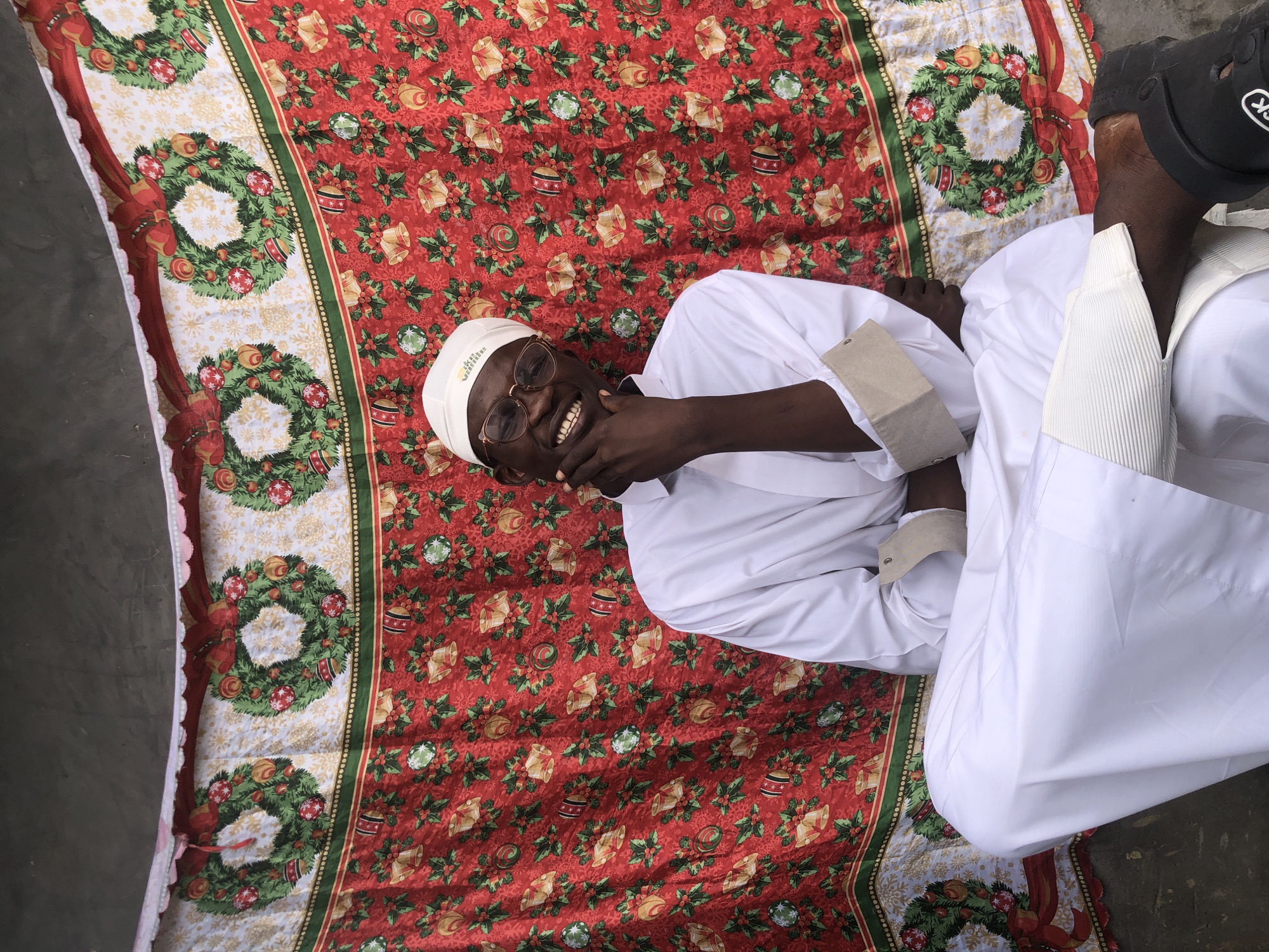 A man wearing a white religious garment and a white cap sits cross-legged in front of a red, green, and white Christmas-themed tapestry. The tapestry features images of wreaths, bells, and holly. The man is smiling and has his hand on his chin. He is wearing glasses. The background appears to be a gray wall. The scene is likely outdoors, possibly in the city of Ado, Nigeria, given the context.