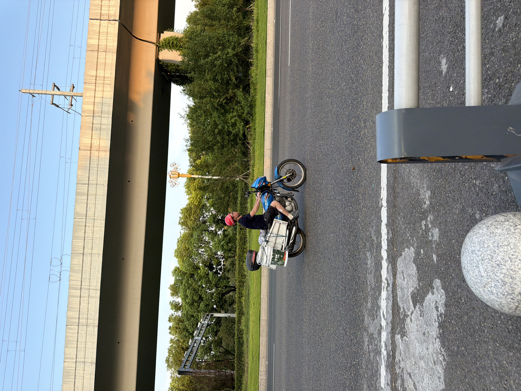 The image shows a person riding a blue motorcycle on a paved road, likely in the city of Jiangjin, China. The rider, wearing a red helmet and a dark t-shirt, is carrying several items strapped to the back of the motorcycle. These items appear to be boxes or containers, some white and some silver, secured with ropes and straps. The motorcycle has a black storage box on the rear.
The setting is outdoors, with a clear blue sky overhead. A large concrete overpass or bridge runs horizontally across the top of the image, with power lines suspended above it. Lush green trees and vegetation line the side of the road, creating a natural backdrop. In the foreground, part of a grey metal barrier with yellow accents is visible, along with a rounded, light-colored bollard. The road surface shows white lane markings and some areas of wear and tear. The lighting suggests it is late afternoon or early evening, with soft shadows cast by the overpass.