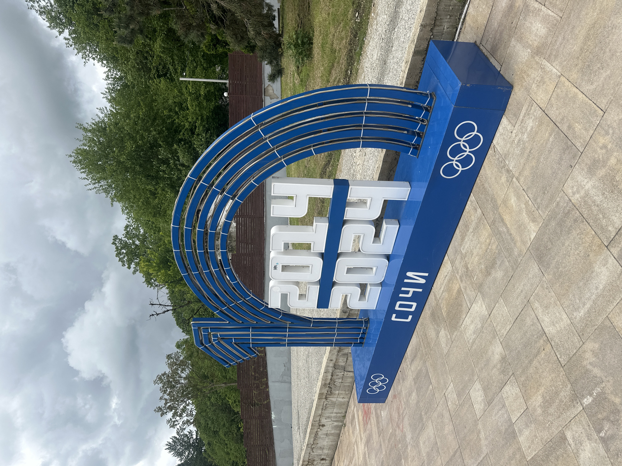The image shows a sign for the 2014 Winter Olympics in Sochi, Russia. The sign is located outdoors in Krasnaya Polyana. It consists of a blue base with the Olympic rings on the front, and white lettering displaying "2014" vertically stacked, along with the name "Сочи" (Sochi) also vertically stacked. Above the lettering, a series of silver-colored, arched metal bars form a semi-circular design. The surrounding environment includes trees, a cloudy sky, and a paved walkway. A wooden fence is also visible in the background. The sign is in a stationary position on the ground.