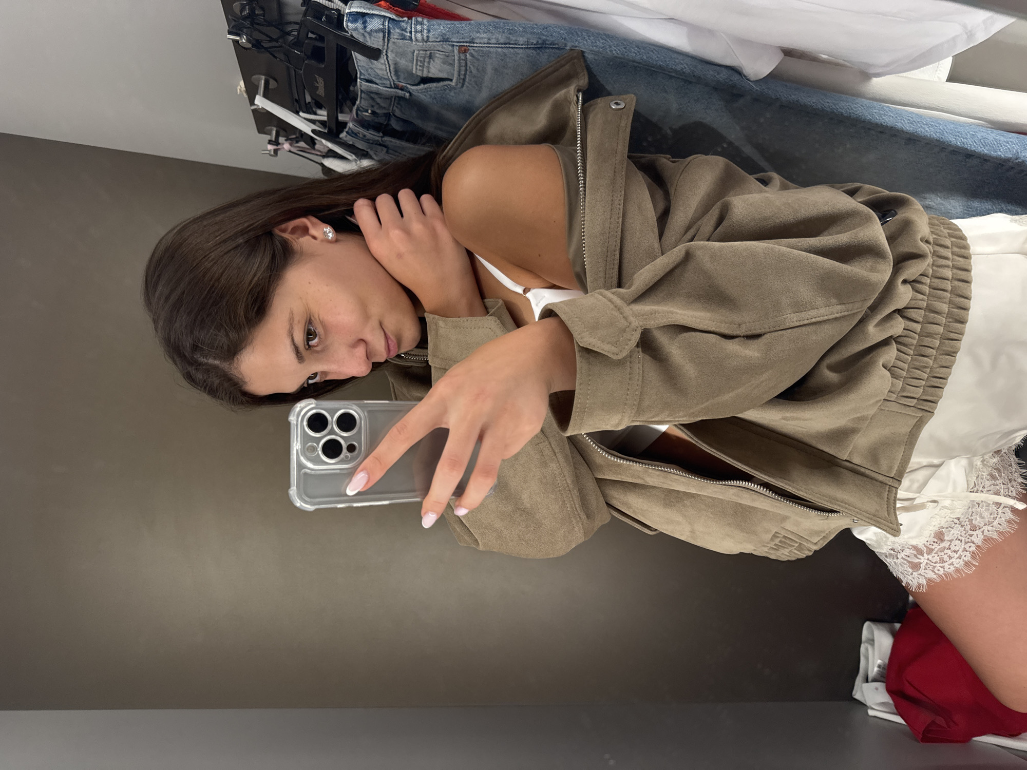 A young woman with dark hair, wearing a taupe suede jacket, white lace shorts, and a white bra, stands in a fitting room and takes a mirror selfie with her iPhone. She is wearing stud earrings and has her hair down. The fitting room has grey walls and a rack of clothes, including jeans and a red garment, are visible behind her. She is holding her phone up to take the picture. The image does not provide enough information to determine the location in the city of Burgas, Bulgaria.