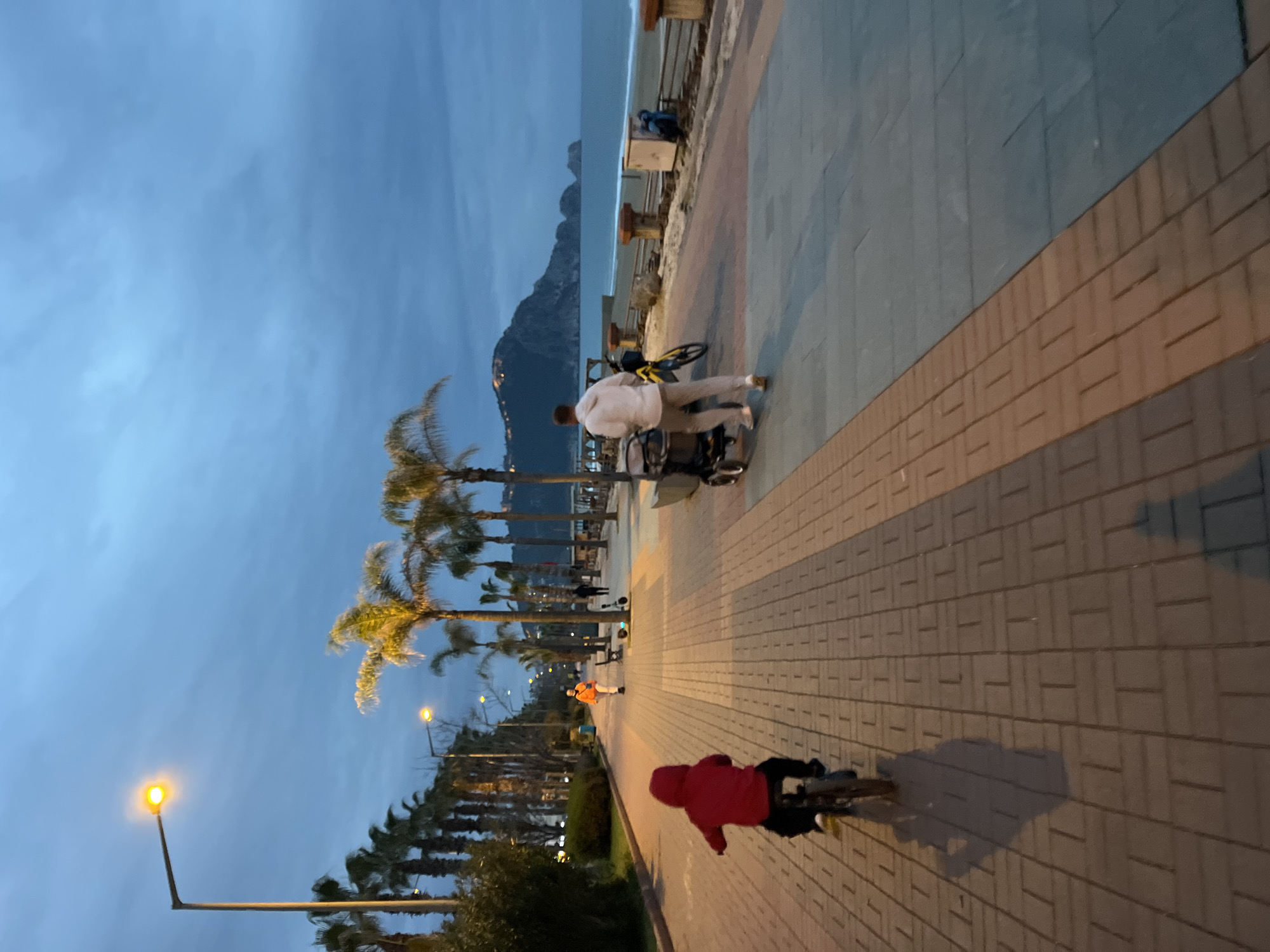Captured in Alanya, Turkey, at dusk, the image showcases a seaside promenade. A cyclist clad in red pedals along the brick-paved walkway, while further ahead, another individual in orange walks. In the background, a person in white stands near palm trees, overlooking the beach. Street lights line the path, and a mountain is visible in the distance. The sky is a muted blue, indicating the early stages of night.