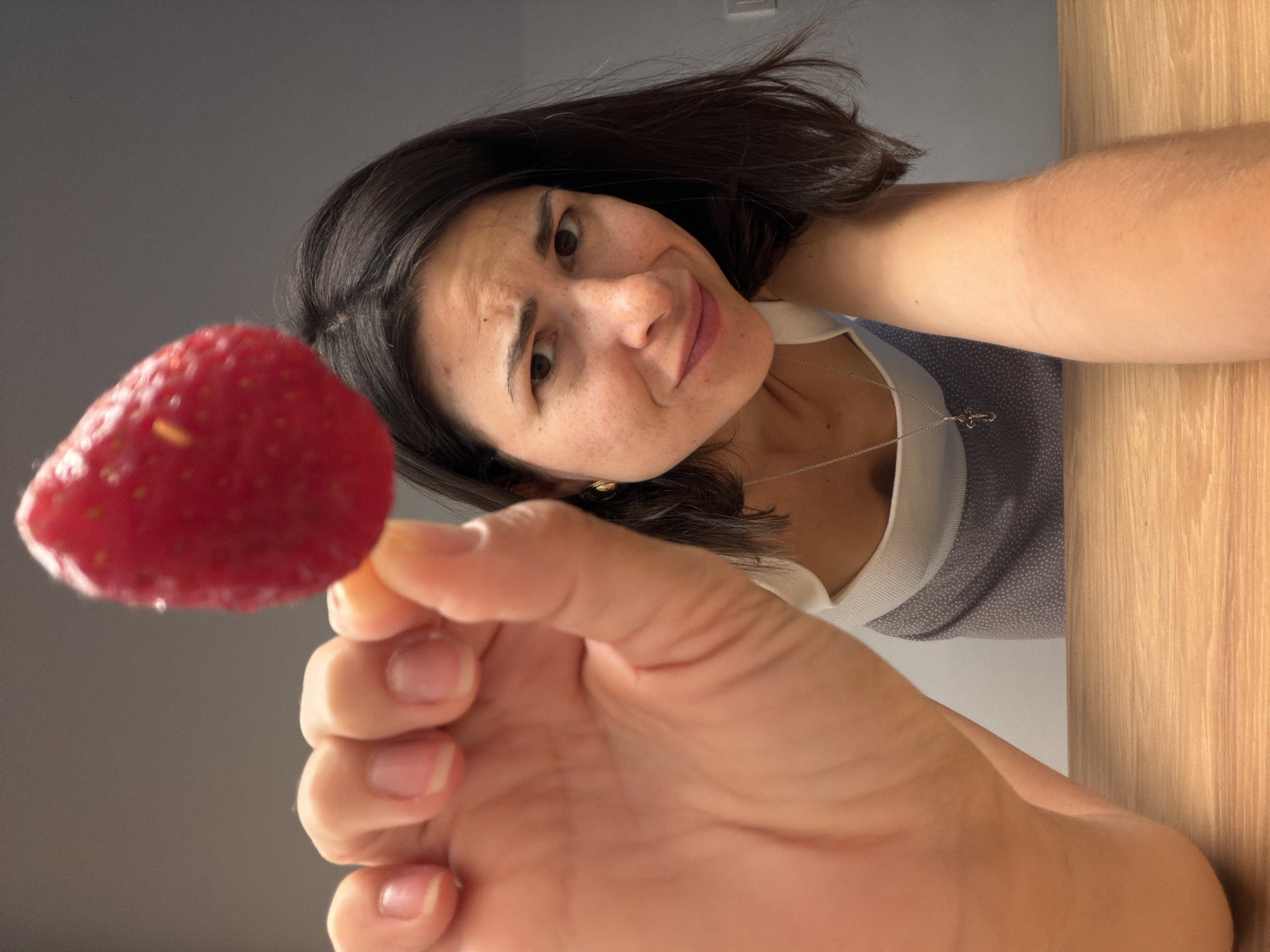 A woman with dark hair and light skin holds a strawberry up to the camera. She is wearing a white and blue top and a silver necklace with a cross pendant. The background is a gray wall and a light brown wooden surface. The woman is looking down at the camera with a slightly amused expression. The scene appears to be indoors. This image does not contain enough information to confirm a location in Budva, Montenegro.