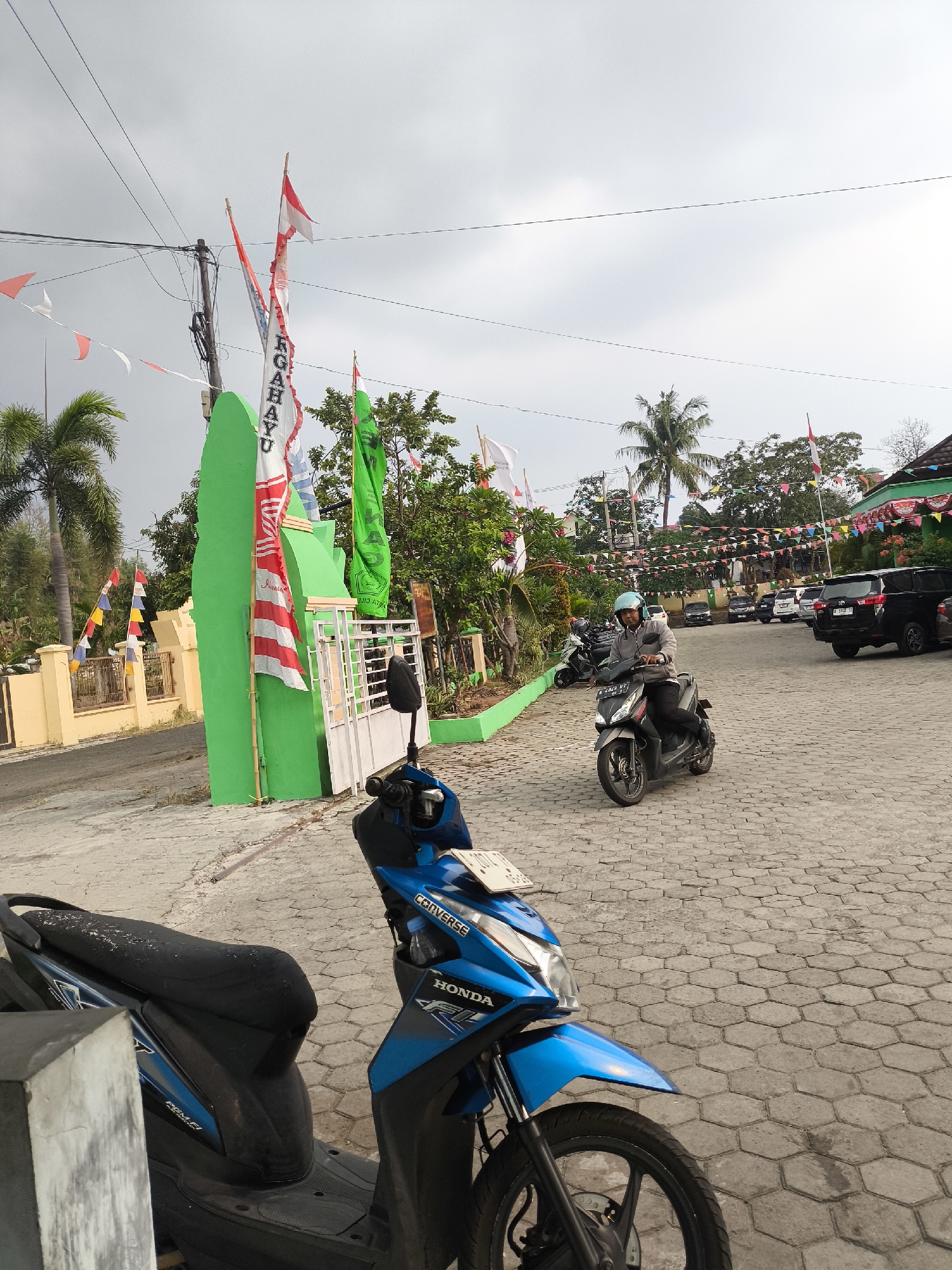 The image depicts a scene in Cilegon, Indonesia, likely in front of a building or complex decorated with national flags and banners. In the foreground, two scooters are parked, one blue and black, and another gray scooter with a rider. The rider, wearing a light blue helmet and a gray jacket, is looking forward as they ride away from the camera on the gray scooter. The ground is paved with hexagonal stone tiles. Behind the scooters, a green gate-like structure is visible, adorned with red and white Indonesian flags and a white banner with text that appears to be in Indonesian. Several trees and more flags are scattered throughout the background. In the distance, cars are parked, and other buildings are partially visible. The sky is overcast. The overall impression is a public area possibly decorated for a local event or celebration.