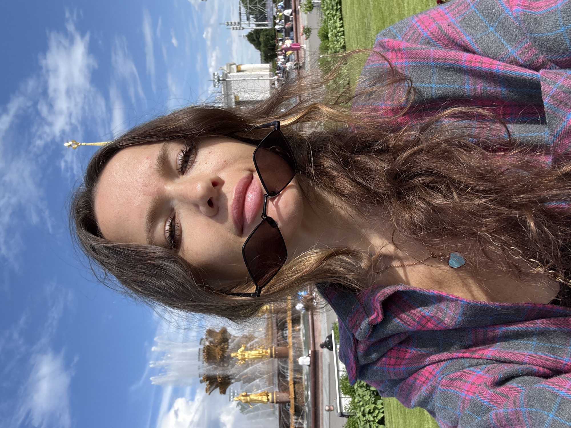 A young woman with long brown hair is taking a selfie outdoors. She is wearing a pink and gray plaid shirt and sunglasses are held in front of her mouth. In the background, a large fountain with golden statues is visible, and a blue sky with scattered clouds is overhead. The fountain and surrounding architecture suggest the location is VDNKh in Moscow, Russia.