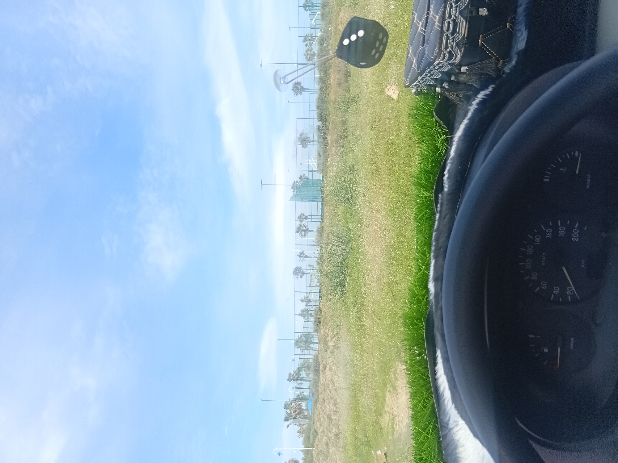 The image, likely taken from inside a vehicle, presents a view of a park or green space in Martil, Morocco. A strip of greenery with trees runs horizontally across the center of the frame. In the foreground, parts of the car's interior are visible, including the dashboard and steering wheel. Outside, the sky is light blue with scattered clouds. A fuzzy dice hangs from the rearview mirror.