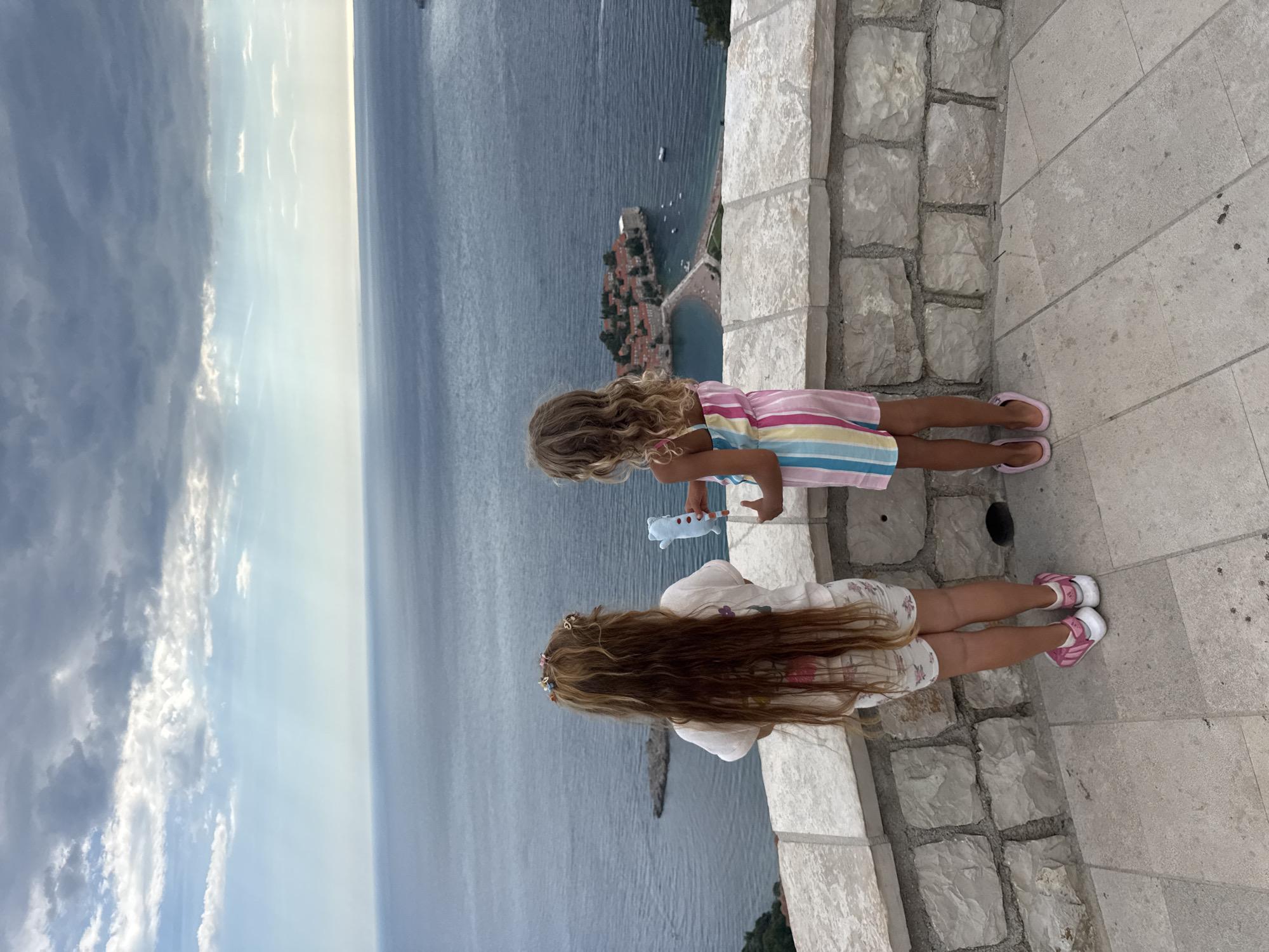In Sveti Stefan, Montenegro, two young girls stand on a stone-tiled terrace overlooking the Adriatic Sea. A grey, cloudy sky with patches of blue stretches across the horizon above the vast expanse of water. The island of Sveti Stefan, a picturesque cluster of red-roofed buildings connected to the mainland by a narrow causeway, is visible in the distance.
The girl on the left, with long, light brown hair adorned with colorful clips, wears a white t-shirt with a floral print and white shorts with a similar pattern. She stands with her back to the camera, holding a light blue object in her right hand. Her feet are clad in pink and white sneakers.
To her right, the second girl, with wavy blonde hair, is dressed in a colorful, striped romper featuring shades of pink, yellow, green, and blue. She also faces away from the viewer, her left hand holding a light blue toy with red polka dots. She wears pink flip-flops.
The girls are positioned behind a stone retaining wall, constructed from large, roughly hewn blocks, which borders the terrace. The wall exhibits a rough texture and variations in color, ranging from beige to grey. The terrace itself is paved with large, rectangular stone tiles, some of which show signs of wear and minor dark stains. The scene conveys a sense of a shared moment of observation, with both children gazing out at the scenic coastal view.