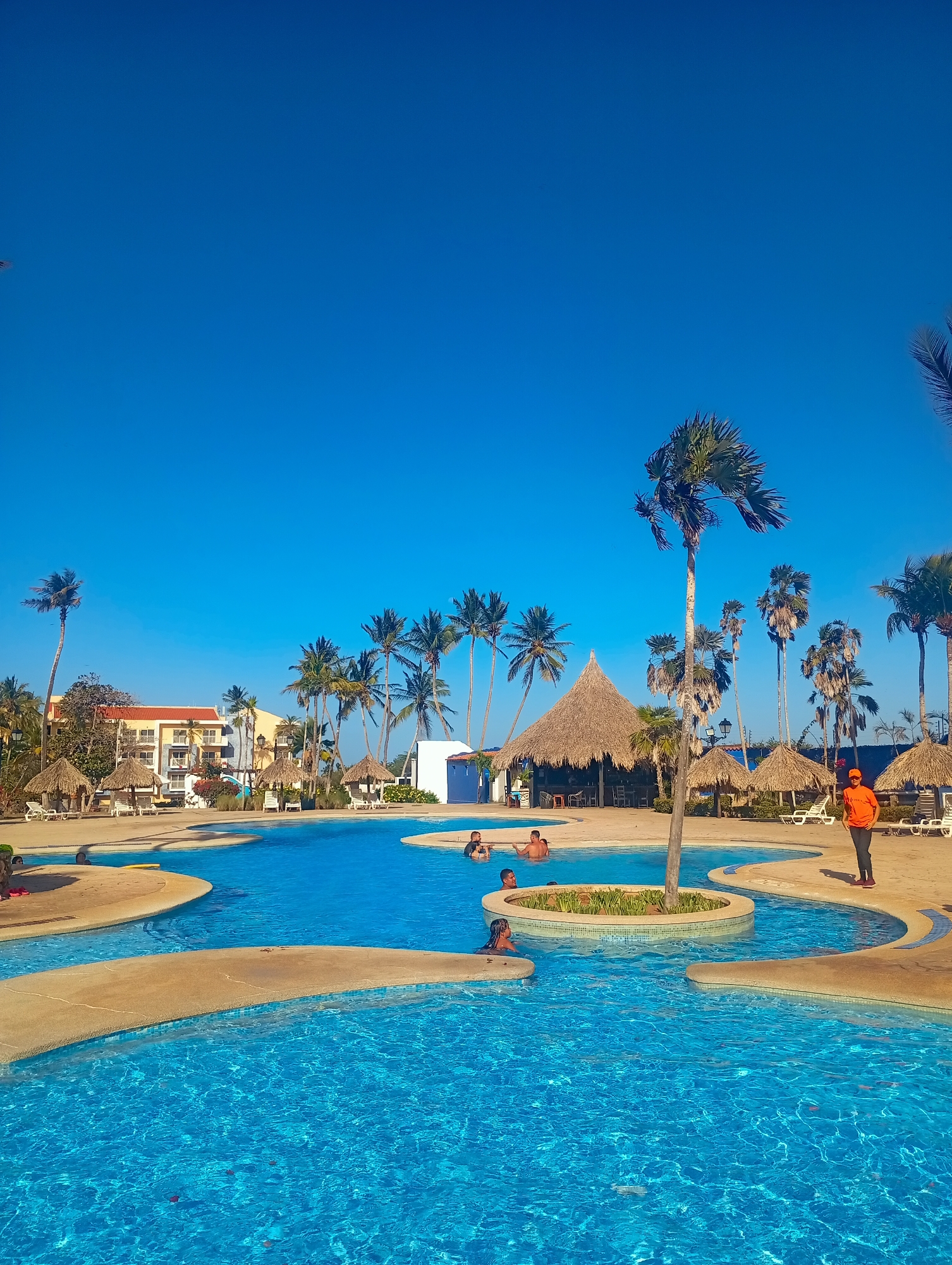 This vibrant outdoor scene captures a resort swimming pool area under a clear, deep blue sky in Playa El Agua, Venezuela. The irregularly shaped pool, filled with brilliant turquoise-blue water, is the central subject, featuring several integrated sandy-colored pathways and small landscaped islands. One prominent tall palm tree stands on an island within the pool.
Several people are enjoying the pool, with individuals seen partially submerged, relaxing or swimming. On the right side of the pool deck, a person wearing an orange t-shirt and dark pants stands, looking into the pool area.
The tropical setting is emphasized by numerous tall palm trees scattered throughout the resort grounds. In the background, a multi-story hotel or resort building with yellow and white facades and red-tiled roofs is visible. Around the pool and between the palm trees, various thatched-roof structures provide shade: a larger hut in the mid-ground appears to be a pool bar or restaurant with interior seating, while several smaller ones function as cabanas or umbrellas shading lounge chairs.
The time of day appears to be late morning or early afternoon, characterized by bright, direct sunlight creating strong reflections on the water and crisp shadows. The weather is perfectly clear and sunny, contributing to an atmosphere of leisure and enjoyment. No visible text is discernible in the image.