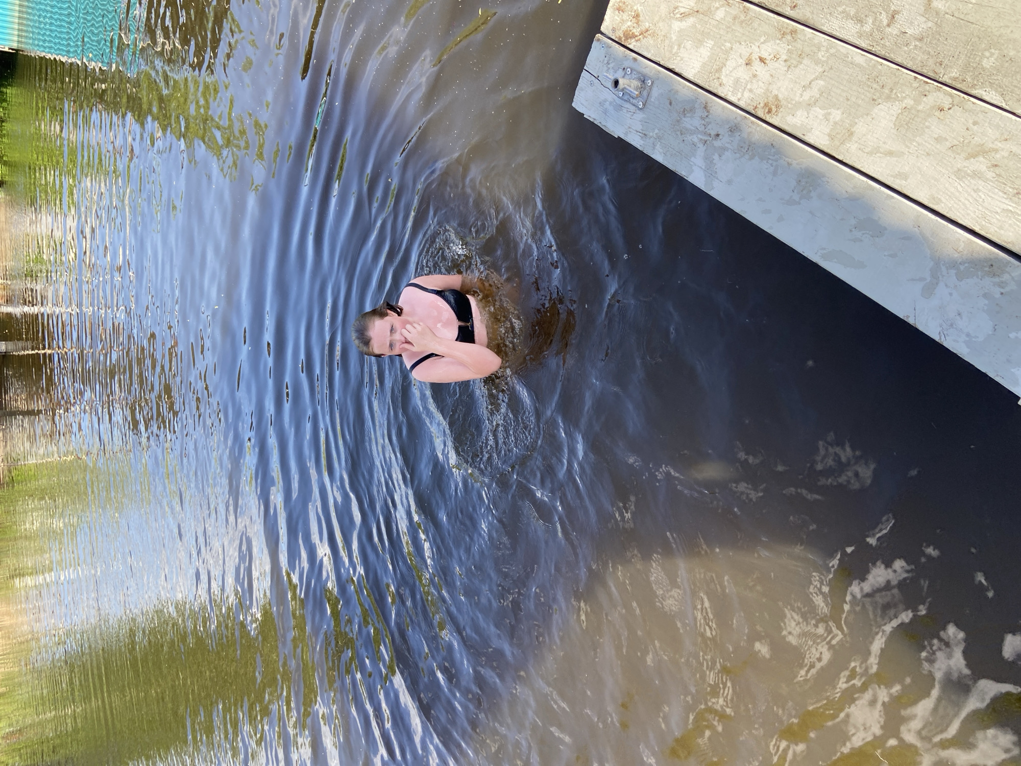 A woman is standing in the water next to a wooden dock. The water surface shows ripples around her. On the left, there are reflections of trees in the water. The location is not definitively identifiable as Chaikovsky, Russia from the visual information provided.