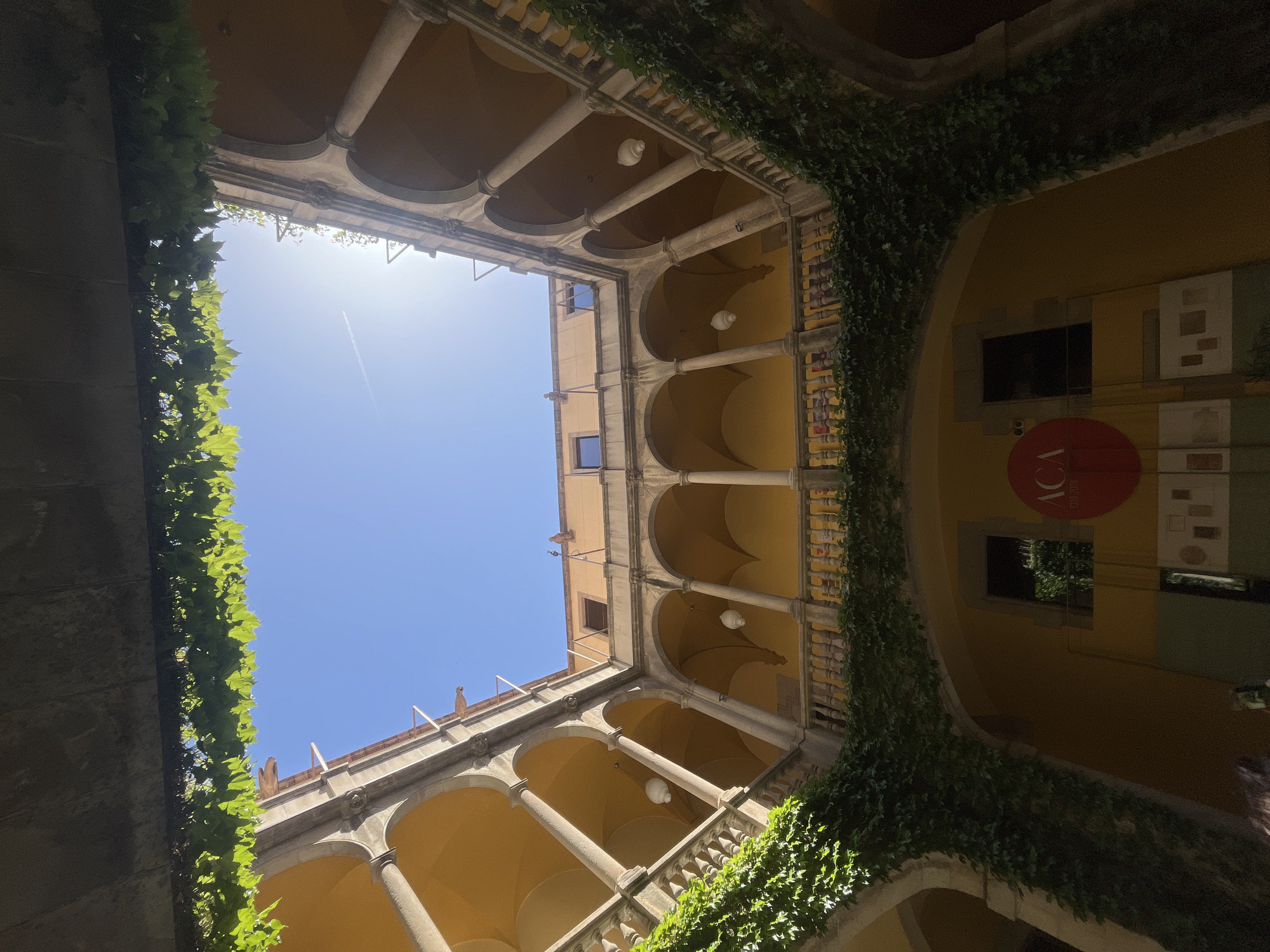 An upward-looking photograph captures the interior of a multi-story building's courtyard or atrium in Barcelona, Spain, revealing a vibrant blue sky overhead. The architecture features classical elements, including a series of arched windows and passageways supported by stone columns and balustrades across multiple levels. The building's walls are painted a warm ochre or yellow, contrasting with the grey stone accents.
Lush green ivy or climbing plants densely cover sections of the balustrades and walls, spilling over the edges. The bright midday sun is visible in the upper left corner of the sky, creating a strong lens flare, and a faint white contrail from an aircraft streaks across the clear blue. Hanging spherical white light fixtures are suspended from the arched ceilings of the lower levels.
On the upper right wall, a distinct red circular sign with white lettering is visible, displaying "ACA" prominently and "1930-2018" in smaller text below it. Several framed pictures or displays are also arranged on this wall. The scene is devoid of people, highlighting the architectural details and the natural light. The bright sunlight suggests a clear, sunny day.