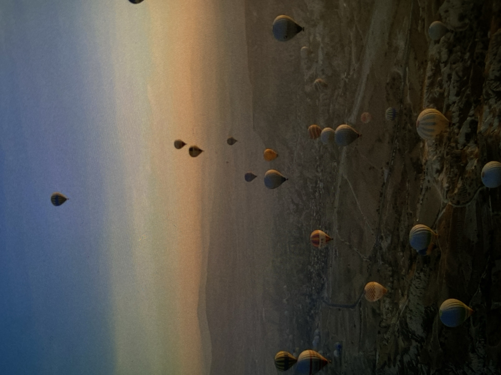 The image displays a panoramic scene filled with numerous hot air balloons soaring over a distinctive landscape, although it is presented in a vertically rotated orientation. When mentally reoriented, with the sky at the top and the ground below, dozens of hot air balloons of varying sizes and colors dot the expansive sky. The balloons exhibit a range of hues including blue, yellow, orange, red, and various striped patterns.
The sky itself presents a serene gradient, transitioning from a light blue at the zenith to warmer shades of soft yellow, orange, and hints of pink closer to the horizon. This atmospheric lighting strongly suggests either a sunrise or sunset, with sunrise being a common time for hot air balloon flights. Below the balloons, the rugged terrain is characterized by unique, eroded rock formations in shades of light brown and grey, indicating an arid or semi-arid environment with a textured, uneven surface.
The activity depicted is a mass hot air balloon flight, likely a popular tourist or leisure event, offering aerial views of the remarkable geological features. The weather appears clear and calm, ideal conditions for ballooning. No people are clearly visible, and there is no discernible text within the image. While the provided location context is Bankya, Bulgaria, the distinctive, eroded rock formations in the image visually strongly resemble landscapes found in regions like Cappadocia, Turkey.