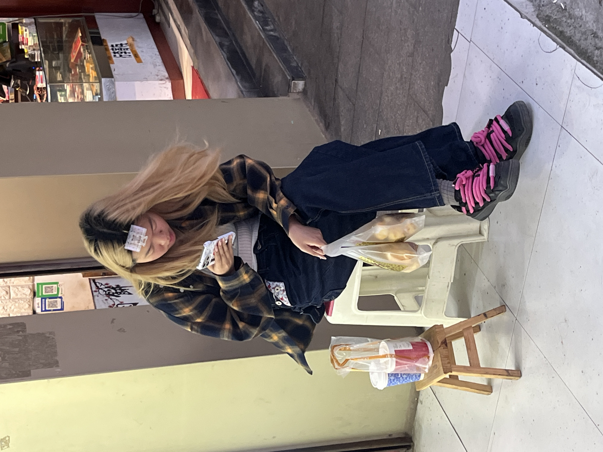 A medium eye-level shot shows a female seated on a white plastic chair, facing left and slightly angled towards the camera, holding a phone with her left hand and an item in her right hand. She wears a black and yellow plaid flannel, a white sweater, and dark blue jeans. Her dark boots have thick pink laces. Clips are visible in her blond hair. The person has purchased a bag of baked goods. A small wooden stool with beverages is placed behind the chair. The location is in the Jiulongpo District of China, as evidenced by the Chinese signs and urban setting. The background includes an entrance to a shop. The floor is tiled.