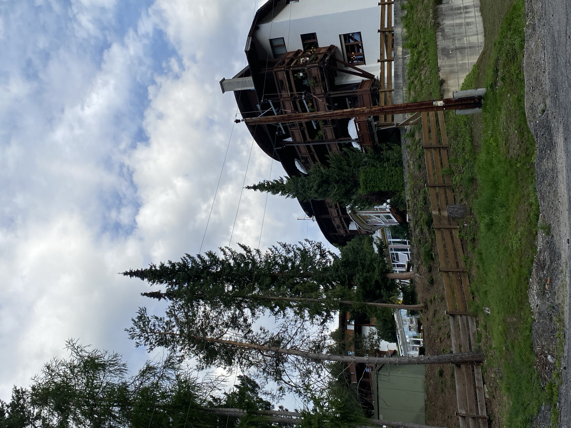 This image captures a view of a building in Reith bei Seefeld, Austria. The building, which appears to be a hotel or a large residence, is multi-story with a white facade and dark wooden balconies. It is situated on a hillside, surrounded by tall evergreen trees. A wooden fence runs along the slope in front of the building, and a dirt and gravel path is visible in the foreground. The sky above is partly cloudy. There are no people visible in the image. The scene is dominated by the architecture of the building and the natural elements of trees and landscape.