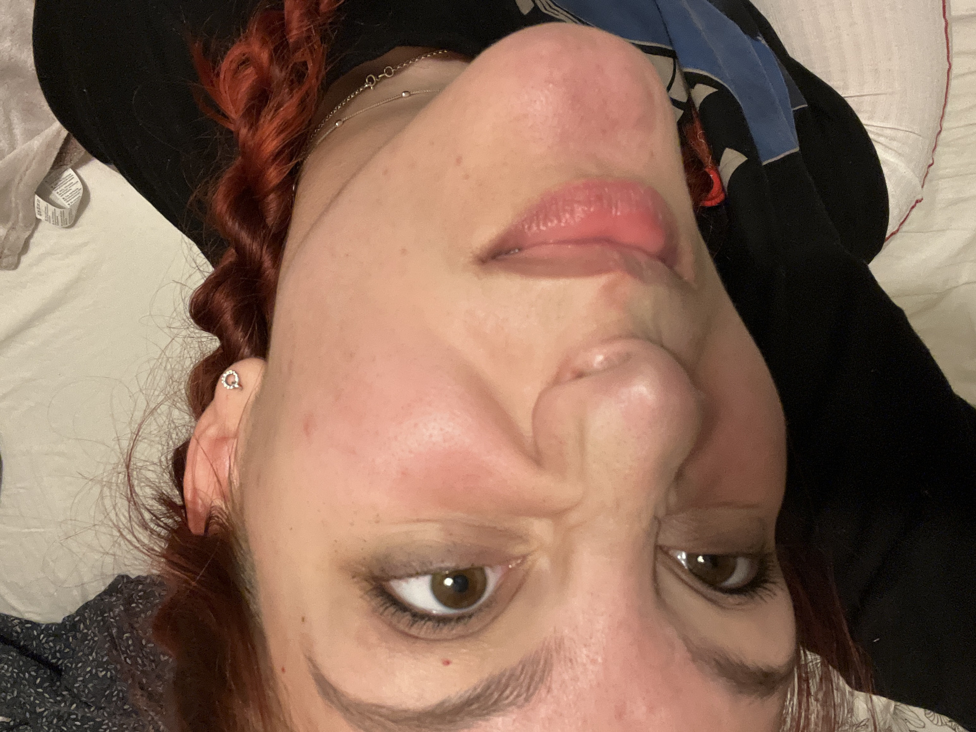 A close-up selfie of a woman with fair skin and reddish-brown curly hair, presumably indoors on a bed. The image is taken from a low angle, looking up at her face. She has brown eyes, defined eyebrows, and slightly parted lips. A silver chain necklace is visible around her neck, and a silver earring is in her left ear. She is wearing a dark top. The background consists of white bedding and a dark patterned garment. There is no indication of the location being in Sofia, Bulgaria, or any specific geographic location.