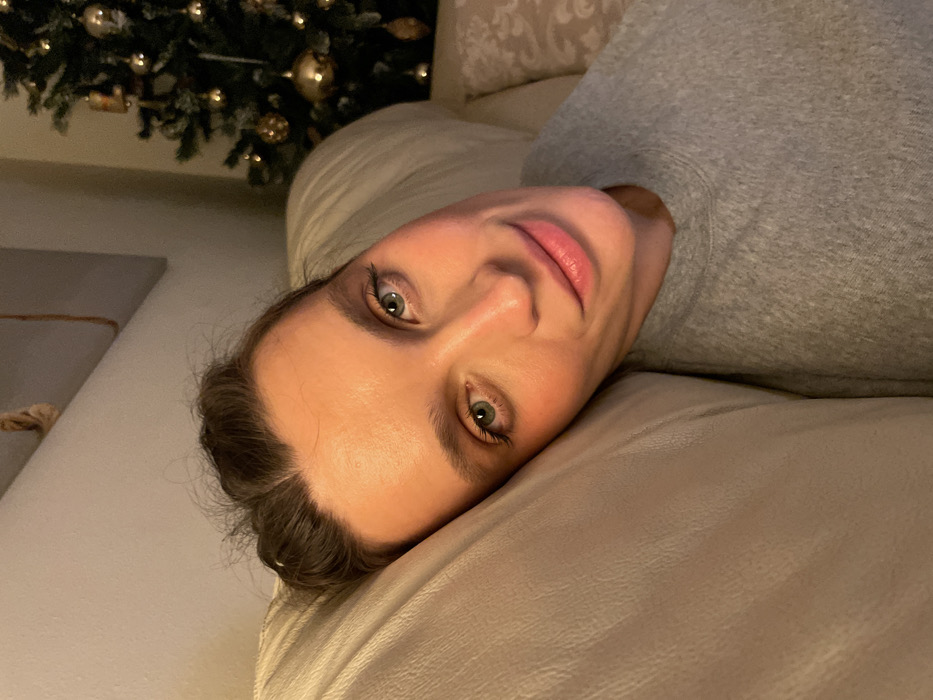 A woman with light skin and green eyes reclines on a beige couch, wearing a gray sweatshirt. Her head is tilted, and she looks directly at the camera with a slight smile. A Christmas tree with gold ornaments is partially visible in the background. The lighting is soft, casting subtle shadows across her face and the couch. The overall scene is relaxed and intimate.
