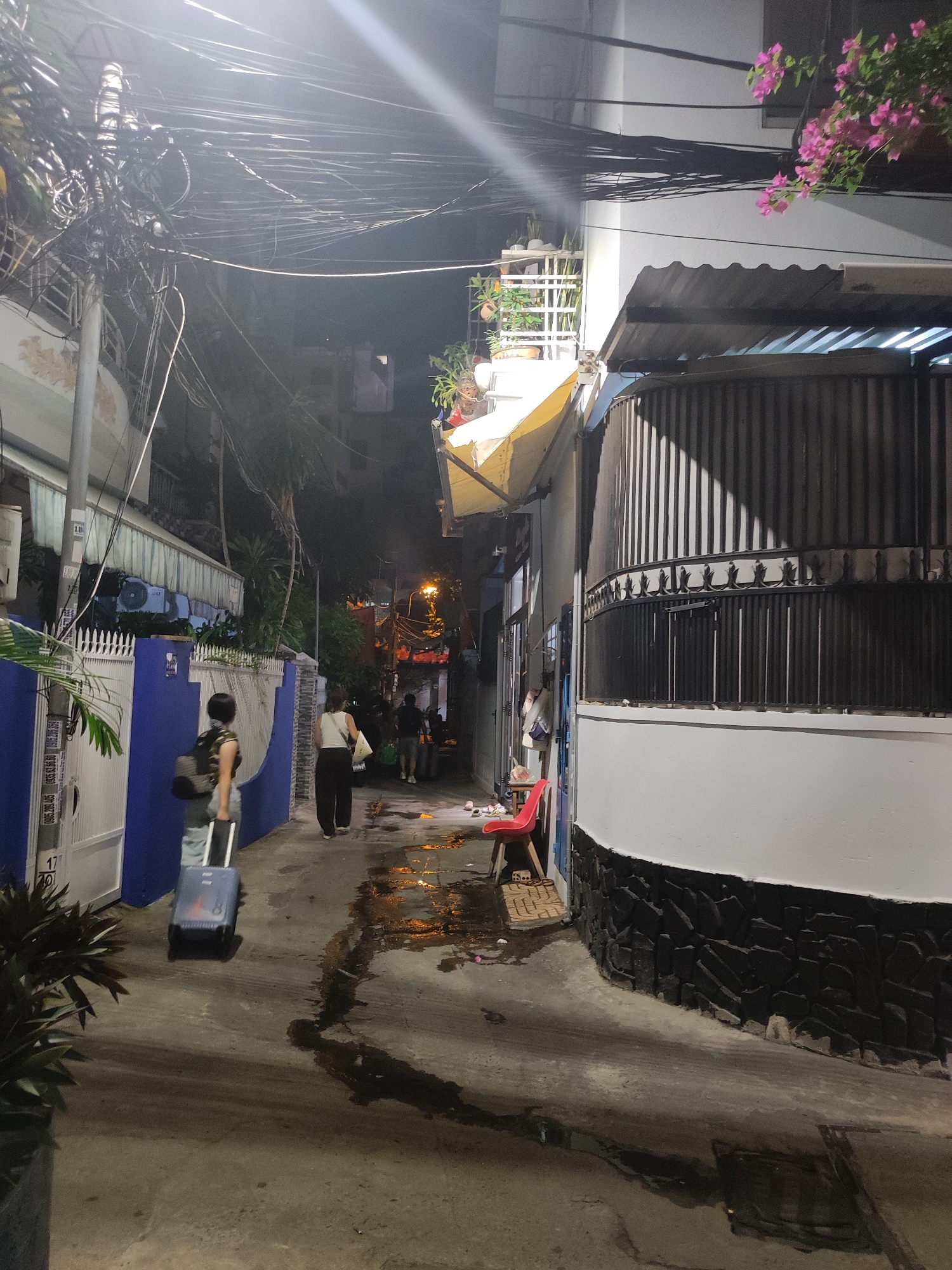 The image depicts a narrow, dimly lit alleyway at night in Nha Trang Ward, Vietnam. Overhead, a dense network of electrical wires is visible against the dark sky, illuminated by a bright light source from the upper left. On the left side of the alley, a blue-and-white fence runs along the property line, with a white gate on the far left. A person is walking away from the camera, pulling a dark-colored suitcase on wheels. The person is wearing a patterned top and dark pants, with a backpack on their shoulders. Further down the alley, two more figures are walking away from the camera.
The right side of the alley is dominated by a building with a white upper section and a lower section clad in dark, irregularly shaped stones. A corrugated metal roof extends from the building, creating a shaded overhang. A dark metal fence with vertical bars and decorative elements runs along the front of this building. To the left of the building, a red plastic chair is visible, placed on a mat.
The ground of the alley is a paved surface that appears wet and reflects the ambient light in several patches, suggesting recent rain or cleaning. Small debris is scattered on the wet ground. In the distance, the alley opens up to reveal more buildings and lights, indicating an urban environment. The overall impression is of a residential or commercial side street at night.